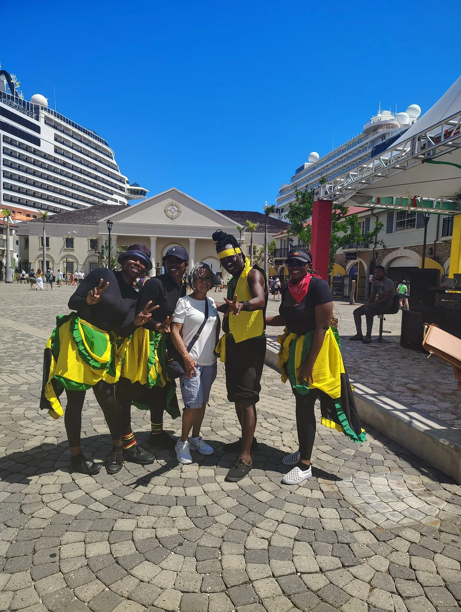 A group of people taking a picture on a sunny day in Jamaica. The dancers are dressed in colorful, coordinated costumes with yellow and green accents, some are making peace signs with a happy tourist in the middle.