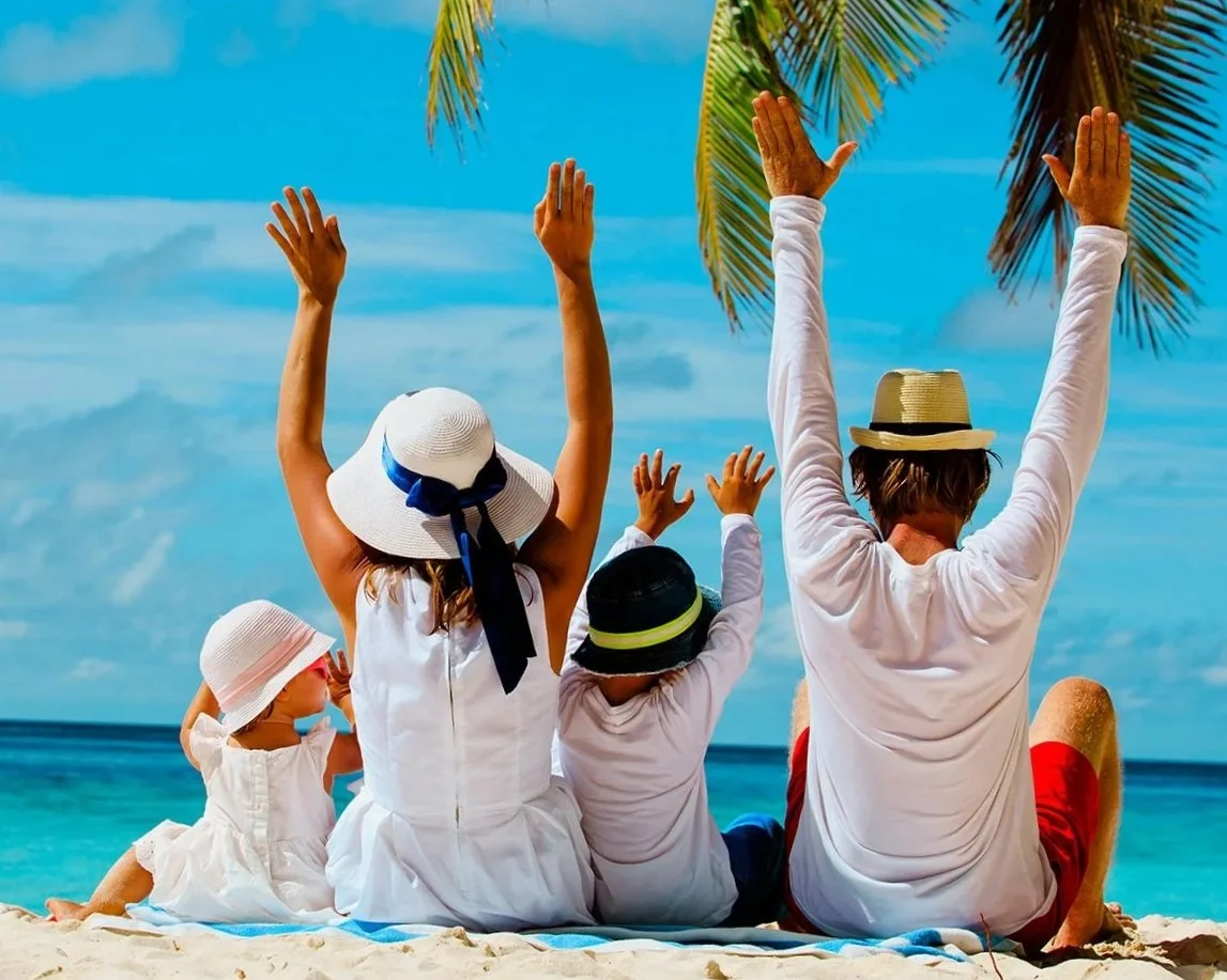 A family sitting on the beach with their backs to the camera, raising their hands in the air, under a palm tree with a blue ocean and sky in the background.