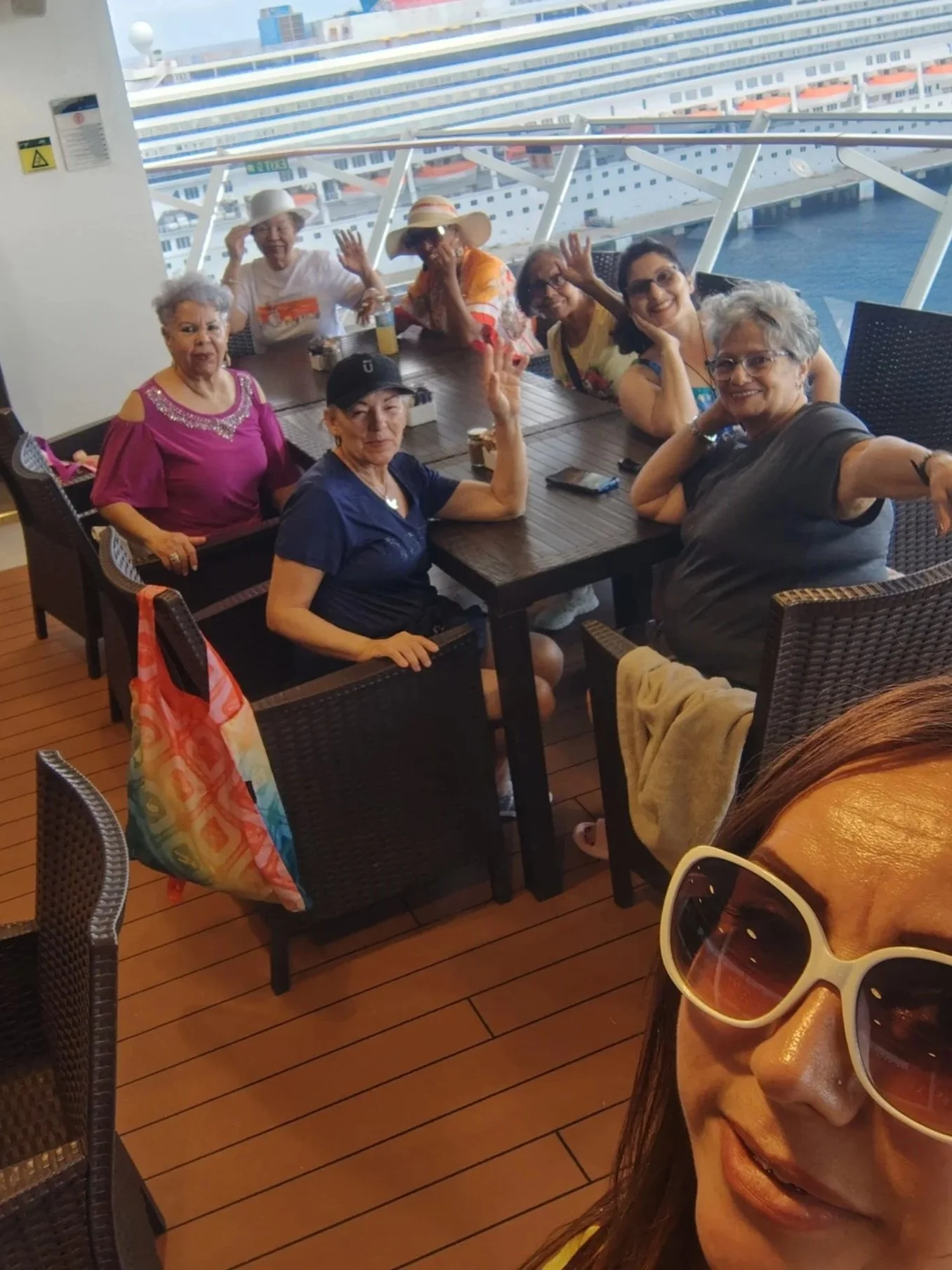 A group of eight happy women sitting around a dining table on a cruise ship, with large windows showing a cruise ship docked outside.