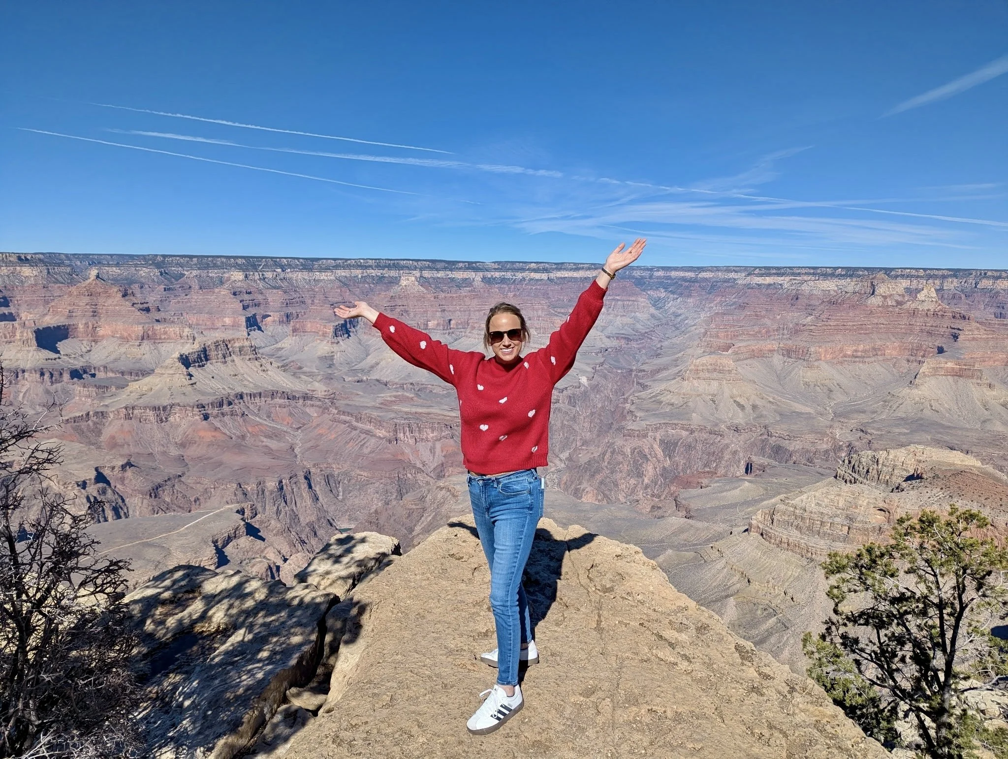 A woman standing on a rock ledge at the Grand Canyon with her arms raised, wearing a red sweater with white heart patterns, blue jeans, white sneakers, and sunglasses, under a blue sky with a few contrails.