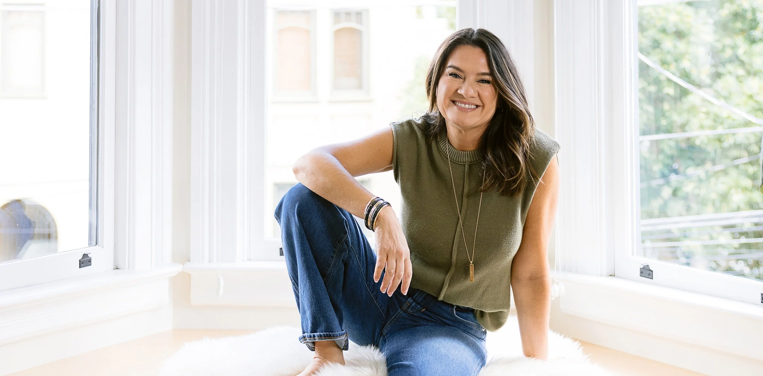 Couples Therapist Andrea Dindinger  with dark brown hair sitting on a white fluffy rug by a large window with a view of green trees outside, wearing a sleeveless green top, blue jeans, and black bracelets.