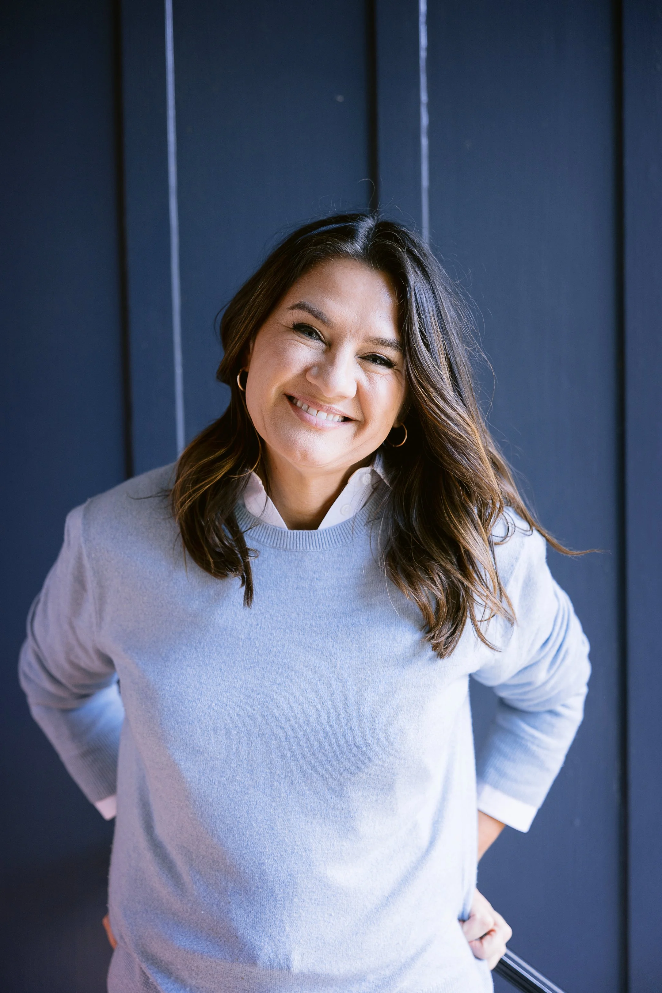 A woman with wavy brown hair, wearing a light gray sweater and white shirt, smiling and standing with hands on hips against a dark blue wall.