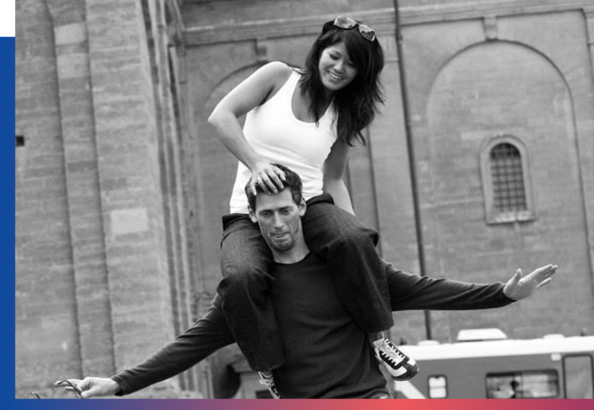 Two people performing balancing acts outdoors with a historic stone building in the background. One person is standing on the other's shoulders, while the other person is lying on the ground, supporting them with an extended arm.