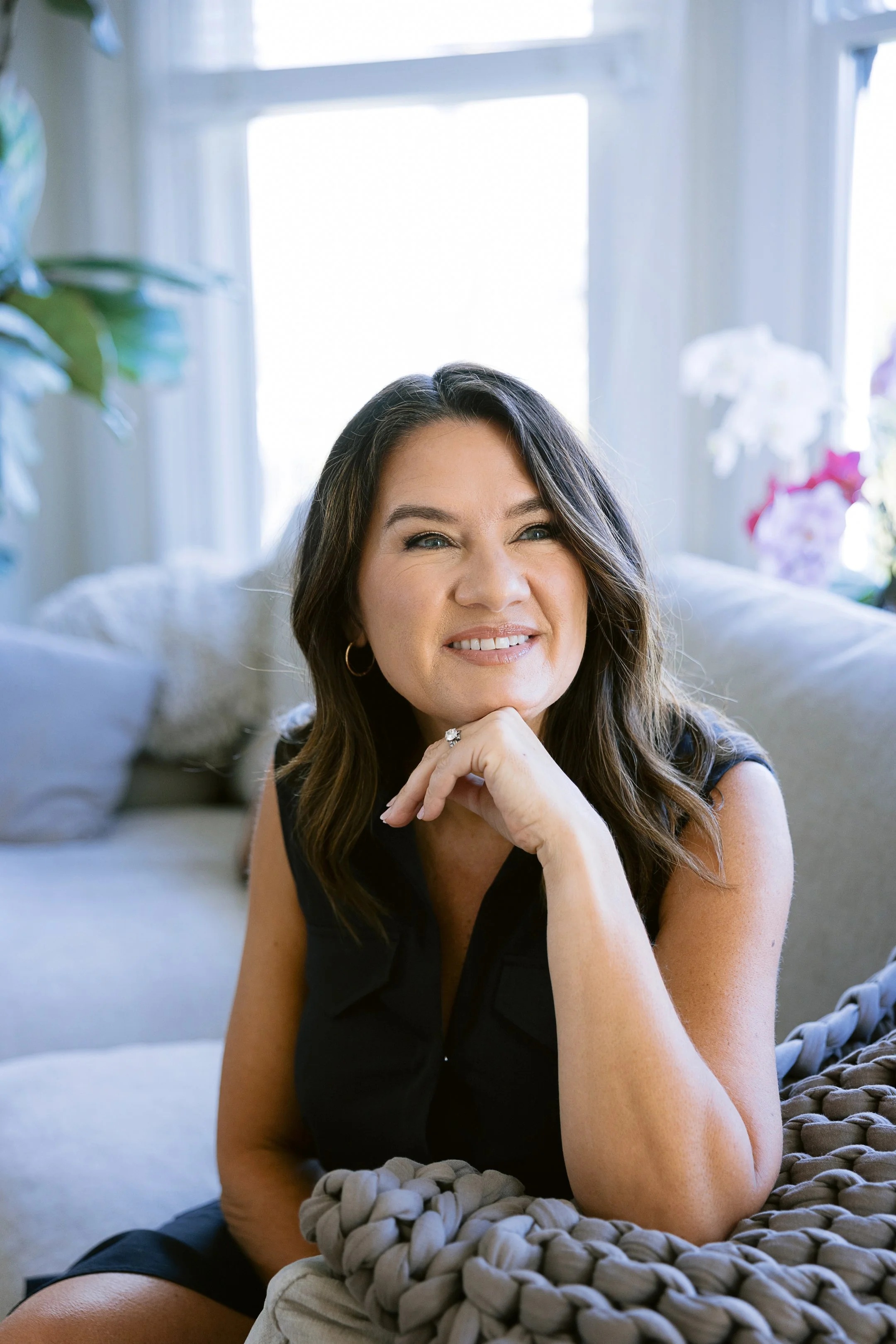 Couples Therapist Andrea Dindinger with long dark hair smiling with her hand under her chin, sitting on a soft knitted blanket in a bright room with large windows and plants around.