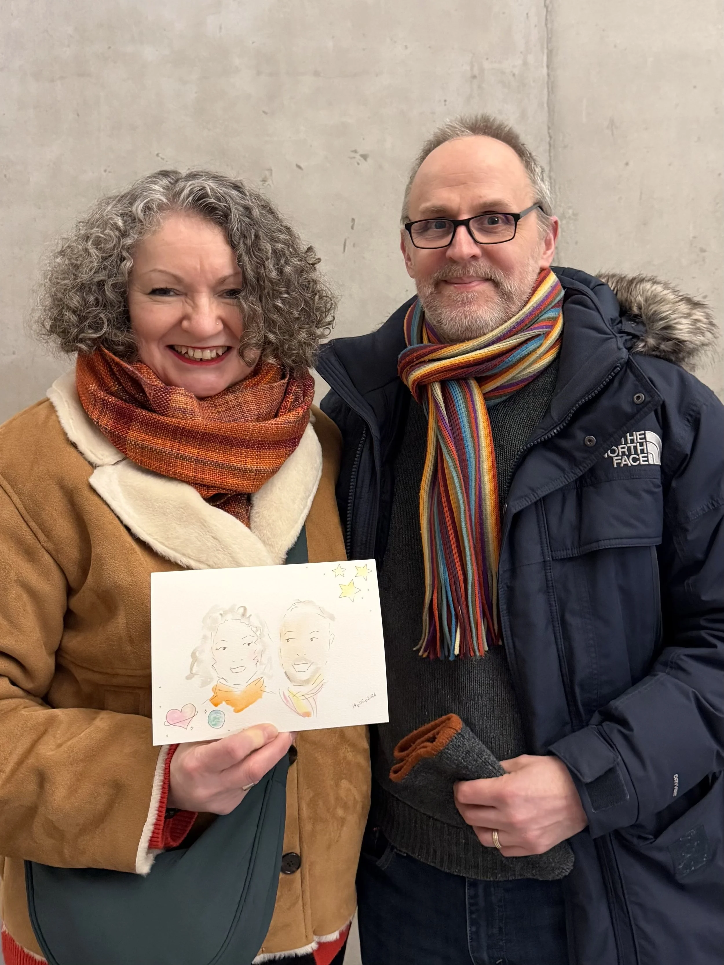 Two smiling people, a woman with curly gray hair wearing a tan jacket and a man with glasses and a beard in a dark jacket with a colorful scarf, holding a piece of paper with a drawing of two faces and some heart and star stickers.