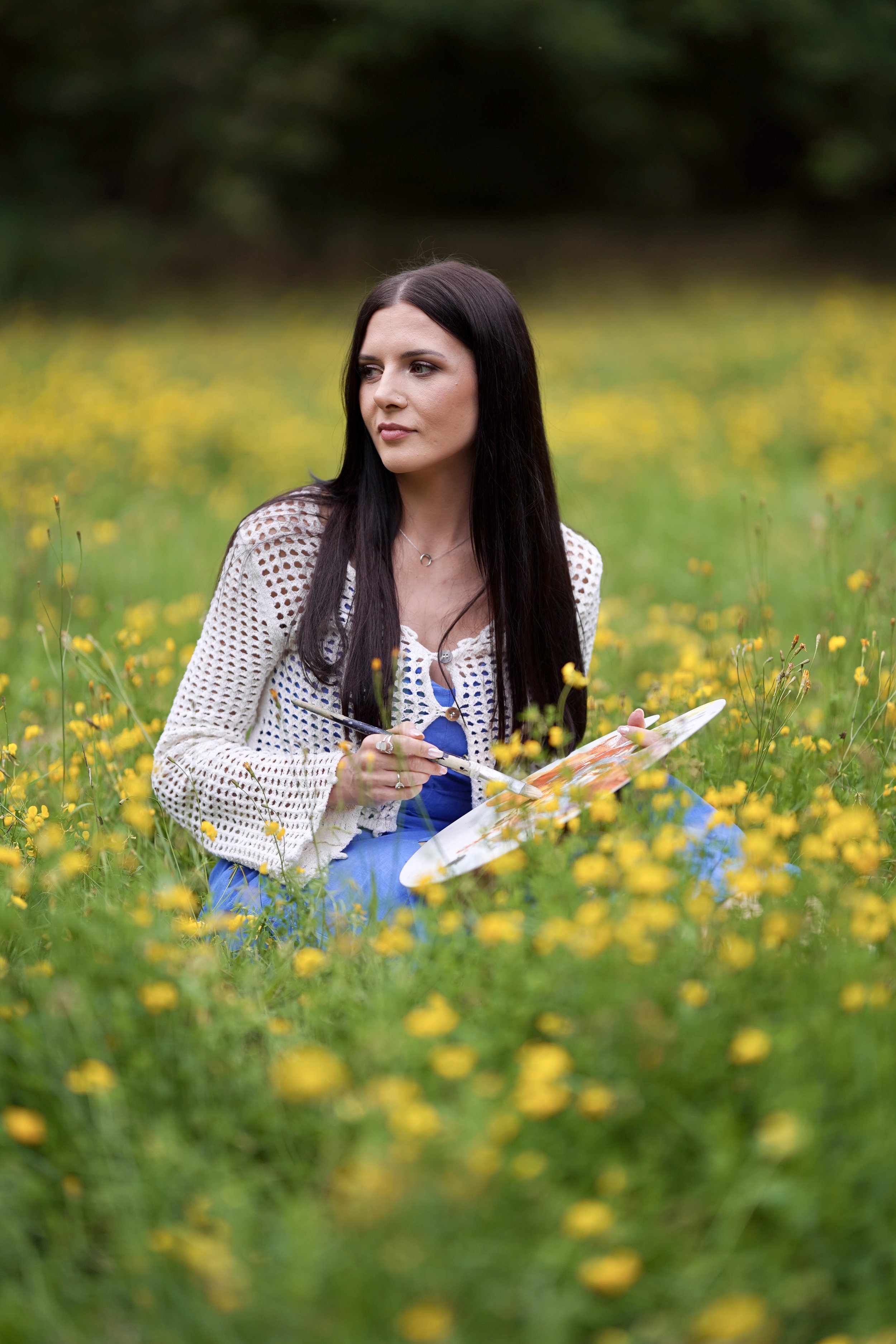 A woman with long dark hair sitting in a yellow flower field, holding a paint palette and brush, looking to the left.
