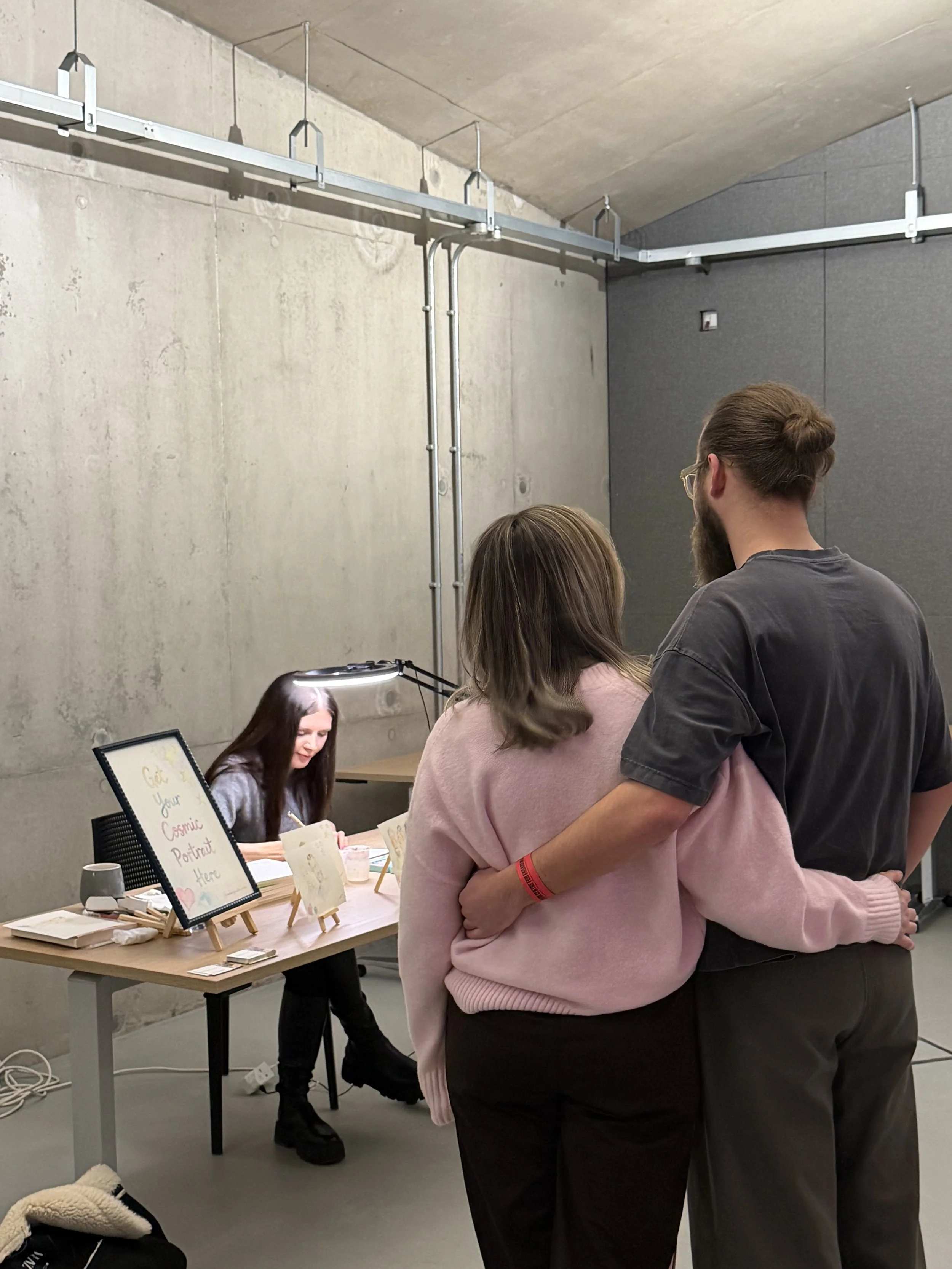 A woman is sitting at a table with a sign that reads "Get your Cosmic Portrait Here" in front of her, while a man and woman stand nearby with their backs turned, inside a room with concrete walls and ceiling.