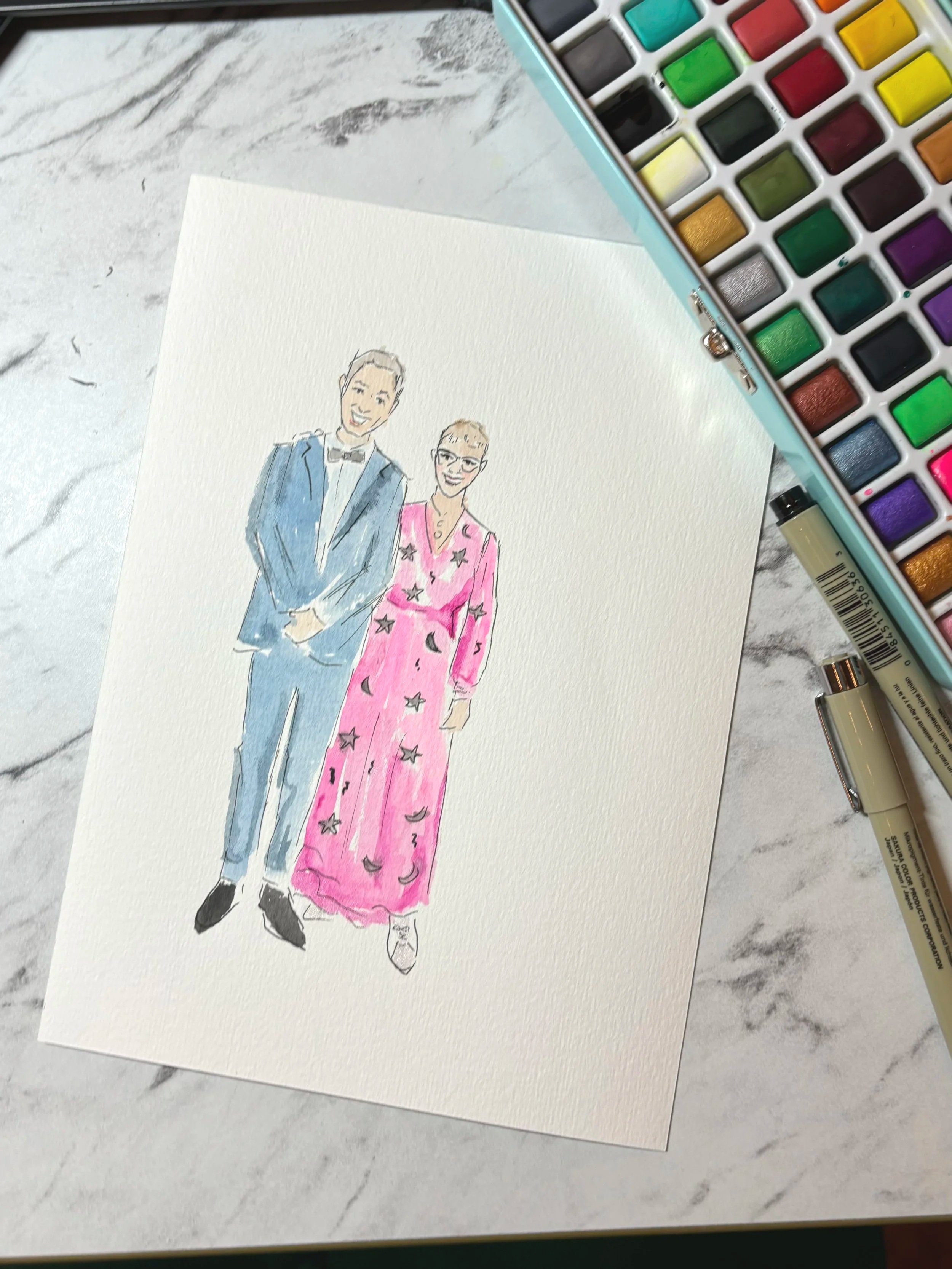A watercolor illustration of a smiling man in a blue suit and black bow tie standing next to a woman in a pink dress with black star patterns. They are on a white sheet of paper on a marble surface, with a set of watercolor paints nearby.