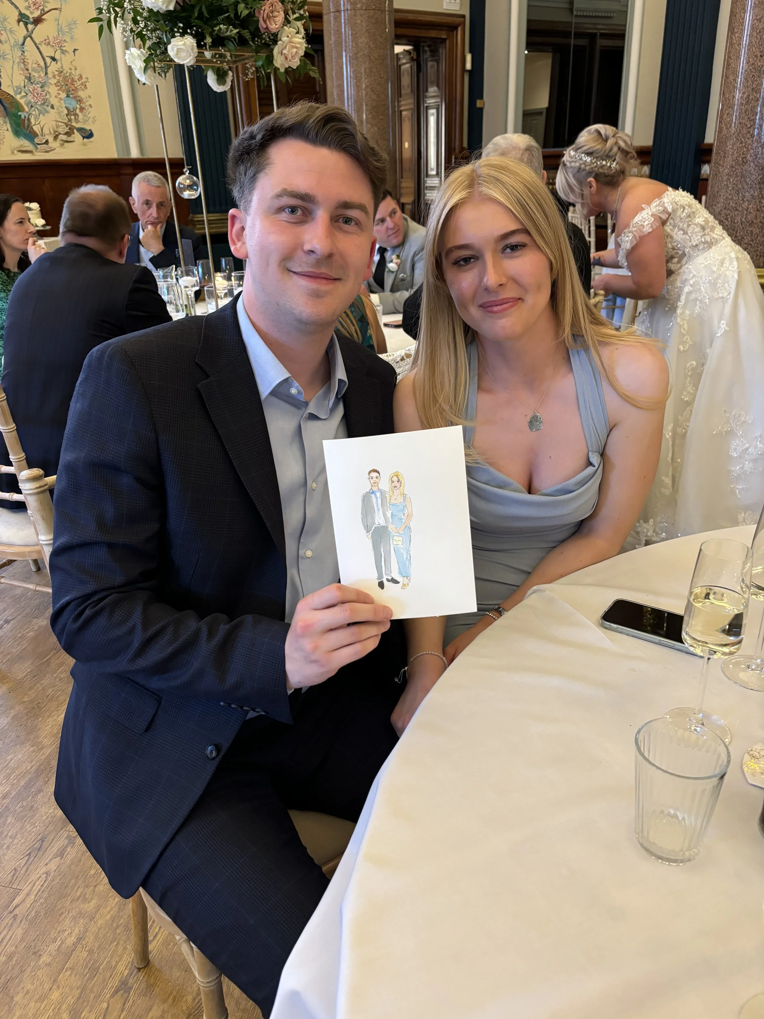A young man in a dark suit and a young woman in a light blue dress sitting at a table during a celebration, holding a card with a drawing of a man and woman. In the background, other guests are seated and a woman in a white wedding dress is standing.