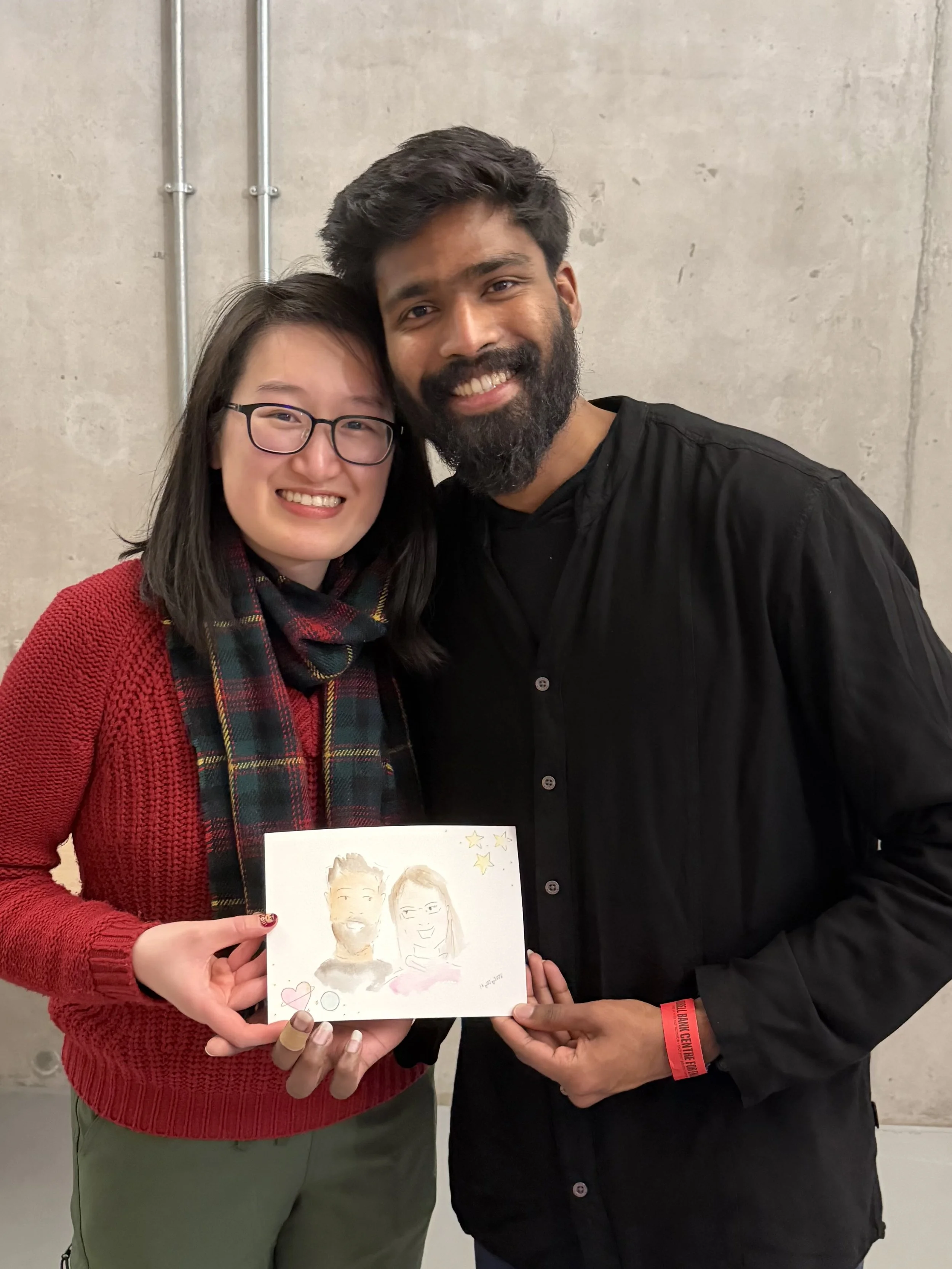 Two people, a woman with glasses and a man with a beard, holding a sketch of themselves together, smiling at the camera against a gray wall background.