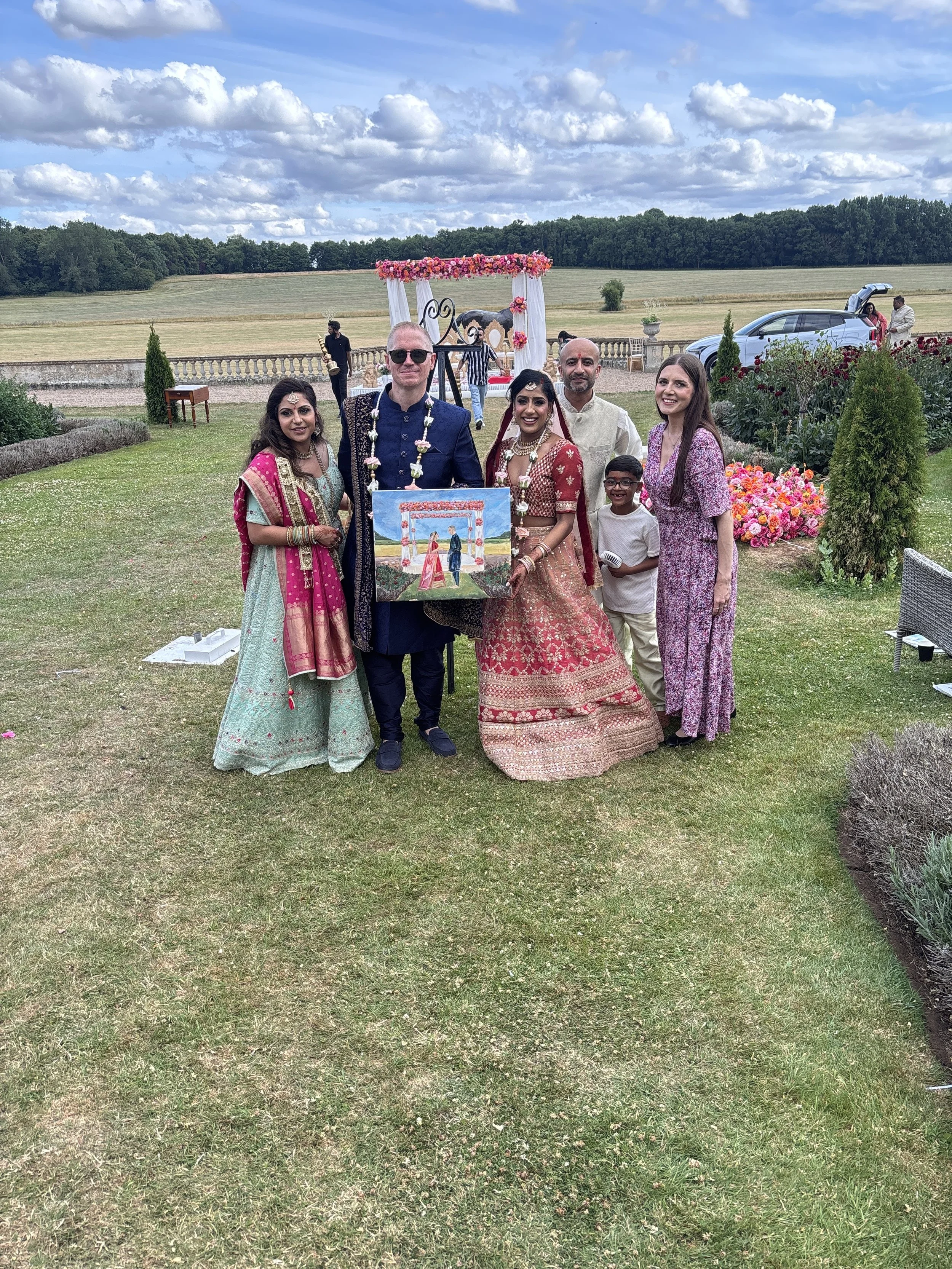 People posing outdoors at a celebration or wedding event, with a decorated arch, a painting, and a scenic countryside background.