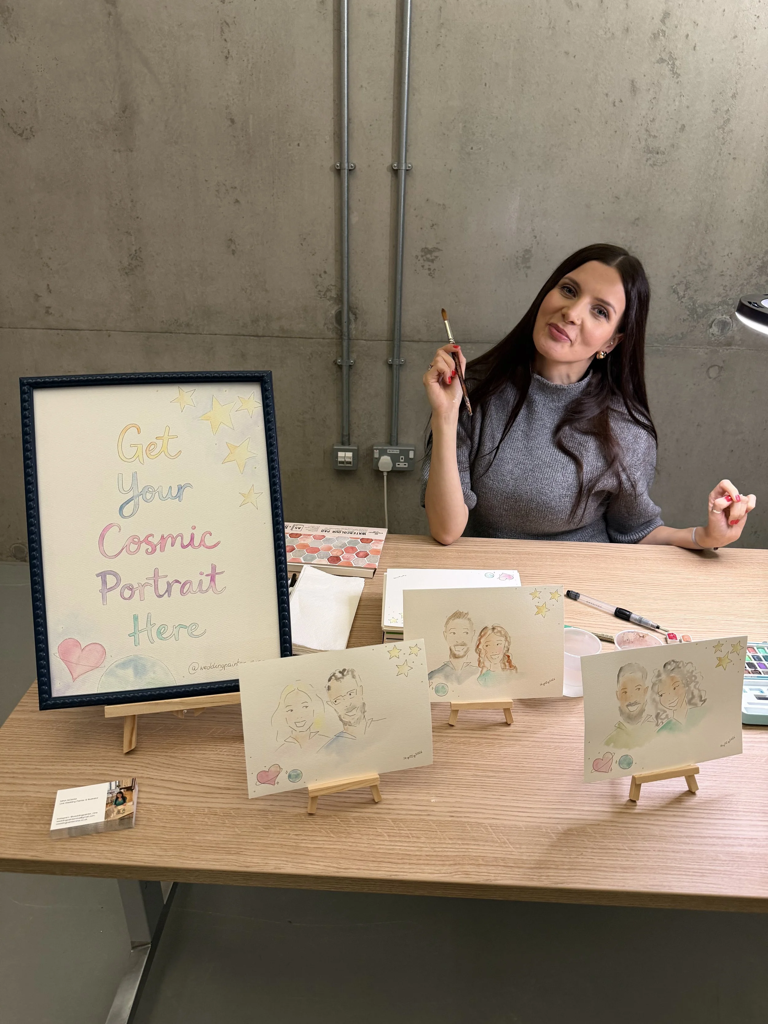 A woman sitting at a table with caricature portraits displayed in front of her. There is a sign that says "Get your Cosmic Portrait" with stars and hearts, and various artistic drawings of people on small easels.