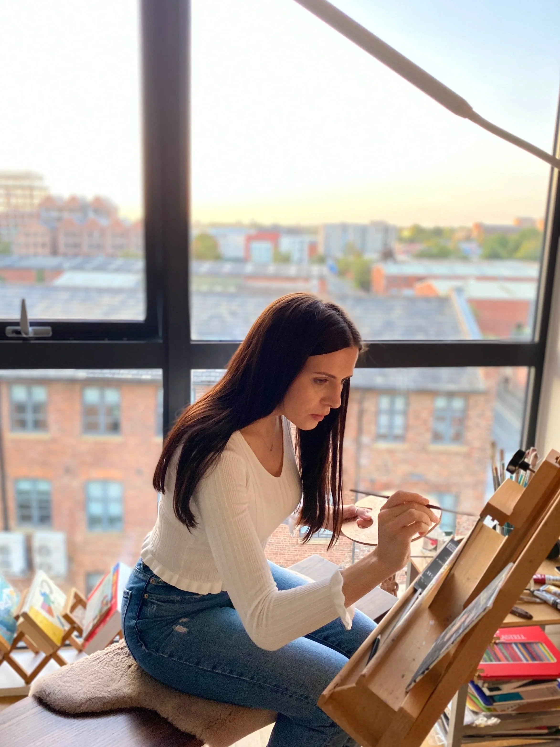 A woman with long dark hair, wearing a white sweater and blue jeans, sitting on a cushioned stool, drawing or writing on a sketchpad placed on an adjustable wooden easel in a bright room with large window overlooking a cityscape with brick buildings and trees.