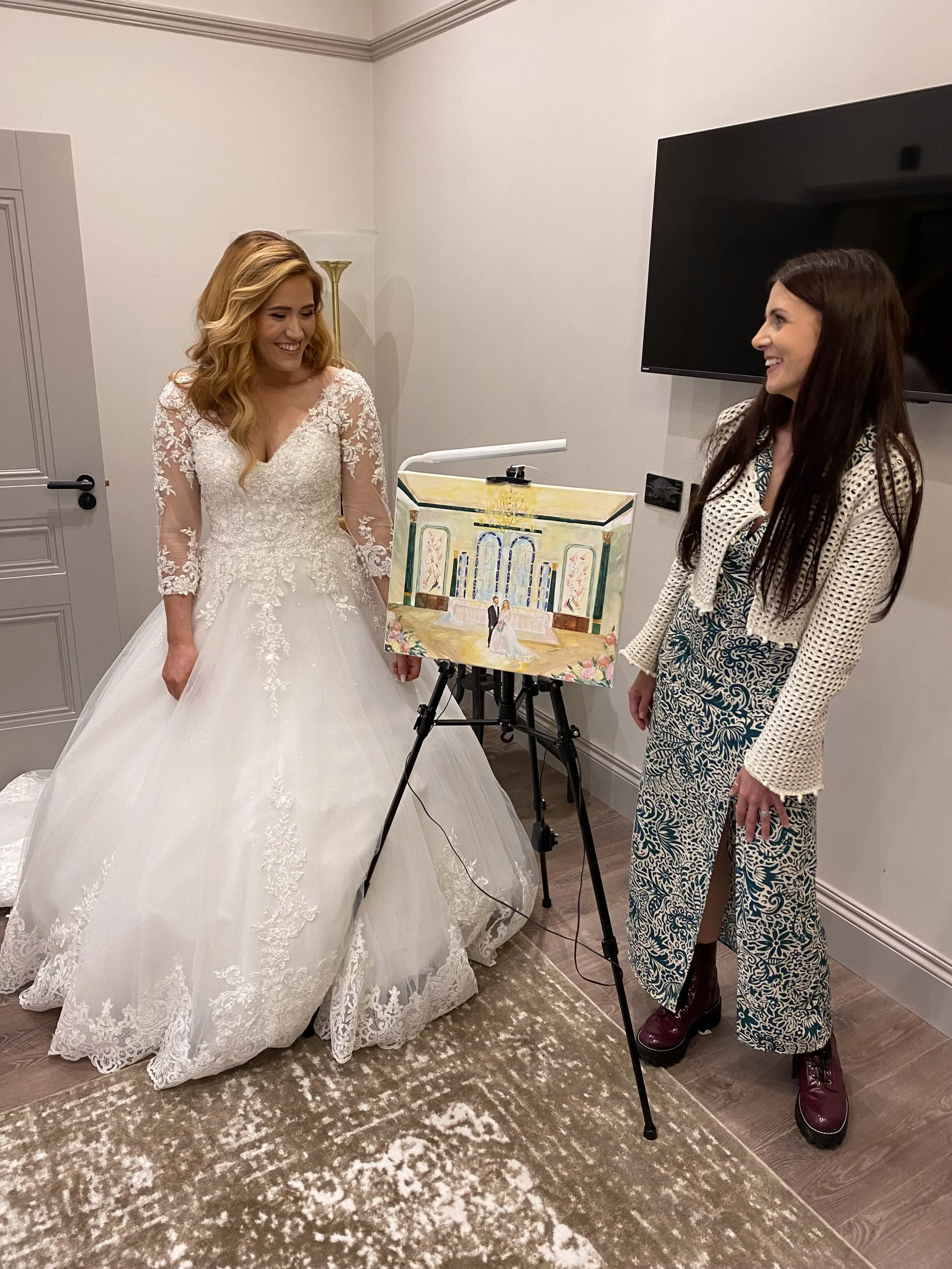 A bride with blonde hair wearing a white wedding dress and a woman with dark hair wearing a patterned jacket and pants, standing inside a room with a large television on the wall. They are smiling at each other, with a painting on an easel between them. The room has white walls, a standing lamp, a door, and a rug on the floor.