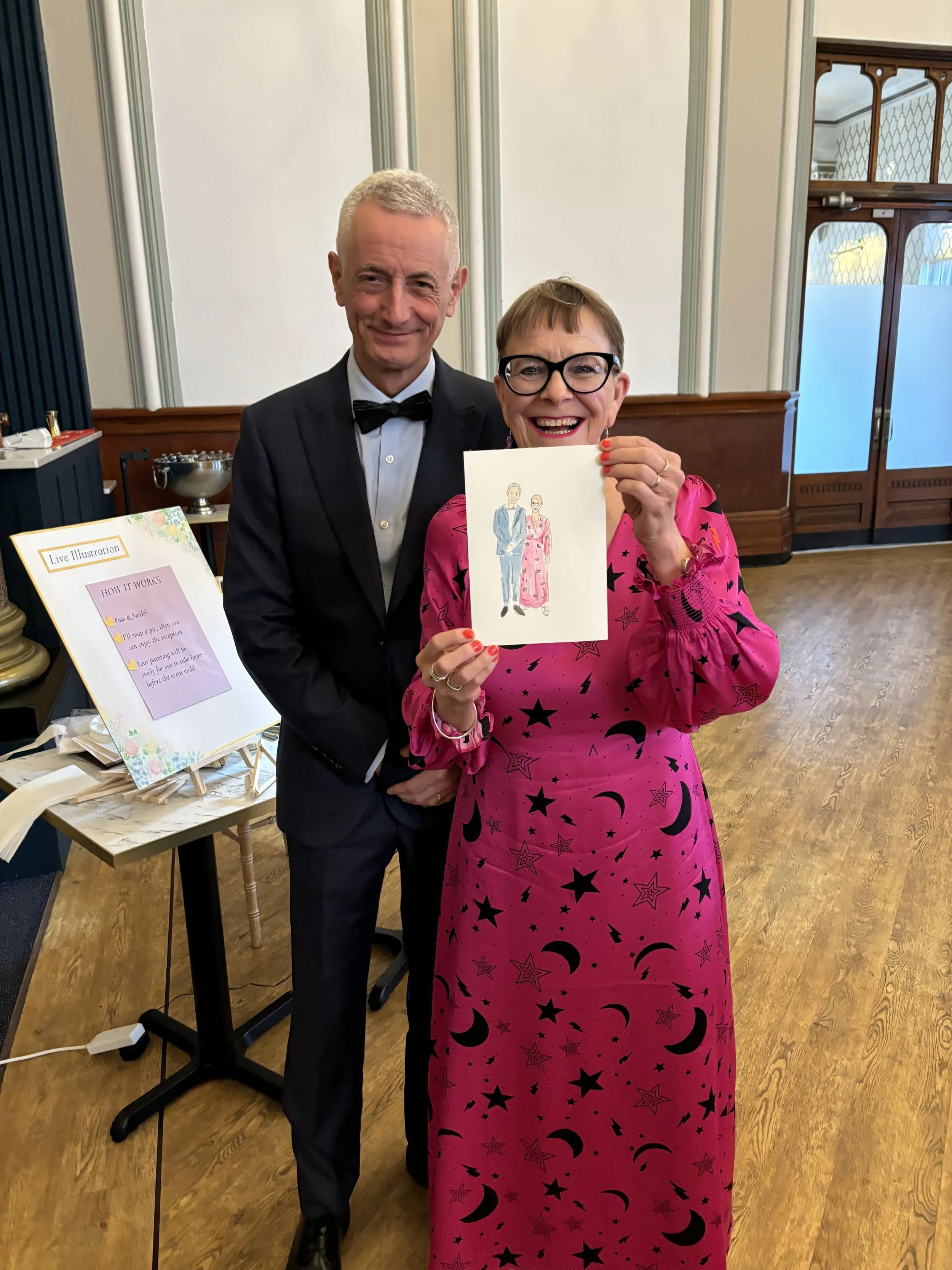 A man in a tuxedo with a bow tie and a woman in a pink dress with black star and moon patterns are smiling and holding a drawing of a man and woman dressed in formal attire. They are indoors with wooden flooring and decorative wall paneling.