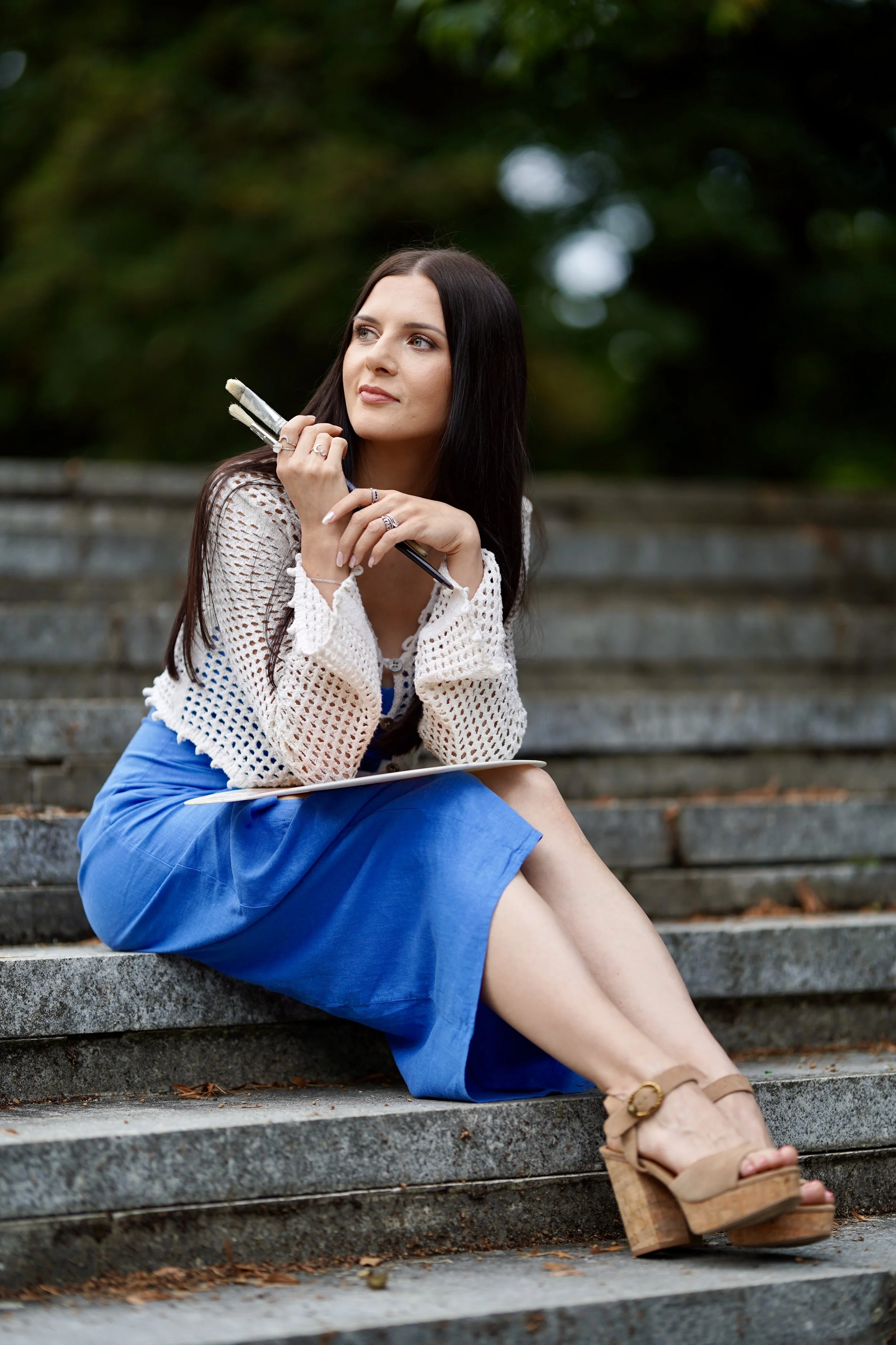 A young woman with long dark hair and fair skin is sitting on outdoor steps, holding paintbrushes and a sketchpad. She is wearing a white crocheted sweater, a blue skirt, a ring, a bracelet, and tan wedge sandals. The background is blurred greenery.