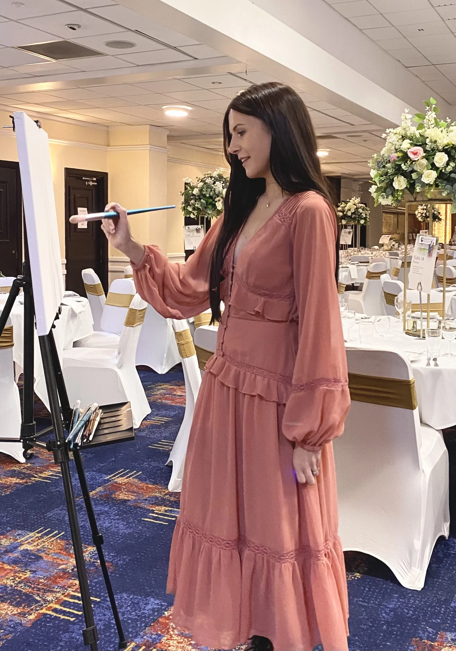 A woman with long dark hair Painting at an event with tables set for a banquet surrounded by large floral arrangements.
