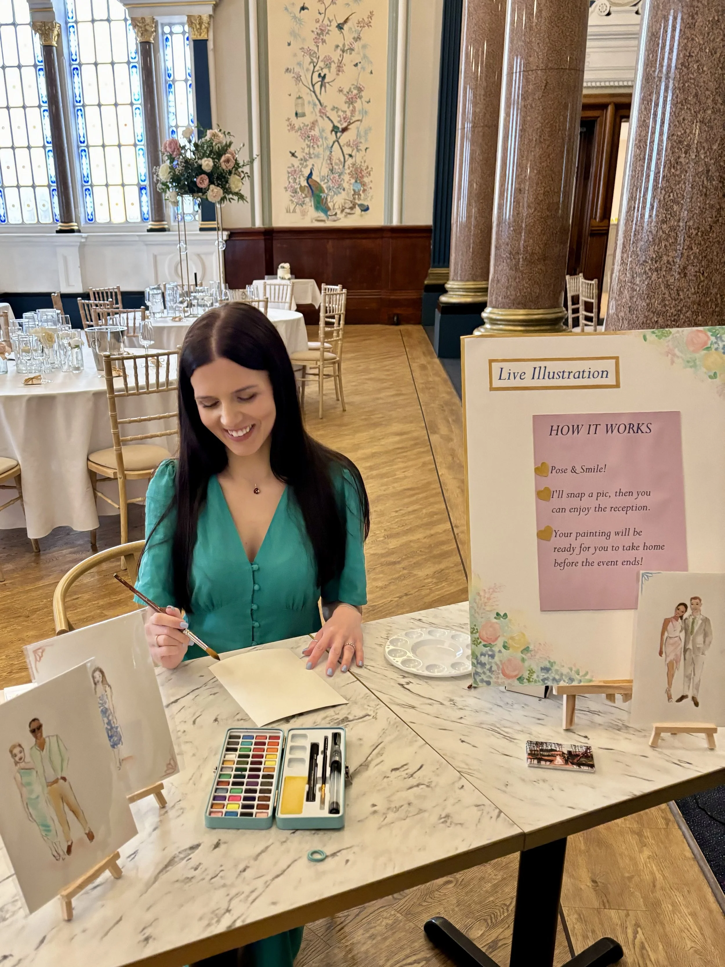 A woman is sitting at a marble table, painting a watercolor portrait of a couple. There are various painting supplies, including watercolor paints, brushes, and paper. In the background, a sign explains the process, and there are elegantly decorated 