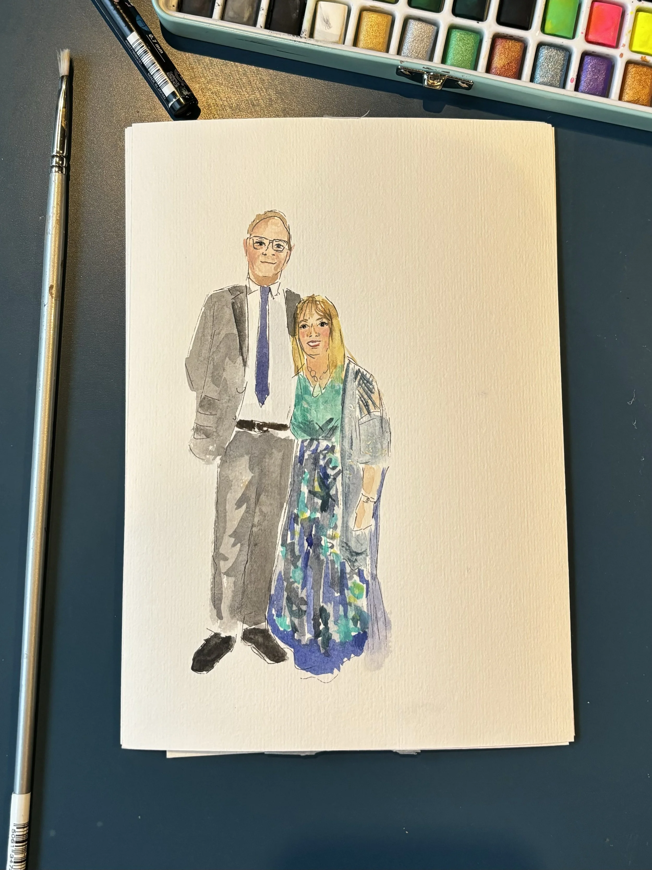 A watercolor painting of a man in a gray suit with glasses and a woman in a colorful dress. The painting is on a white sheet of textured paper, surrounded by watercolor supplies and brushes on a dark table.