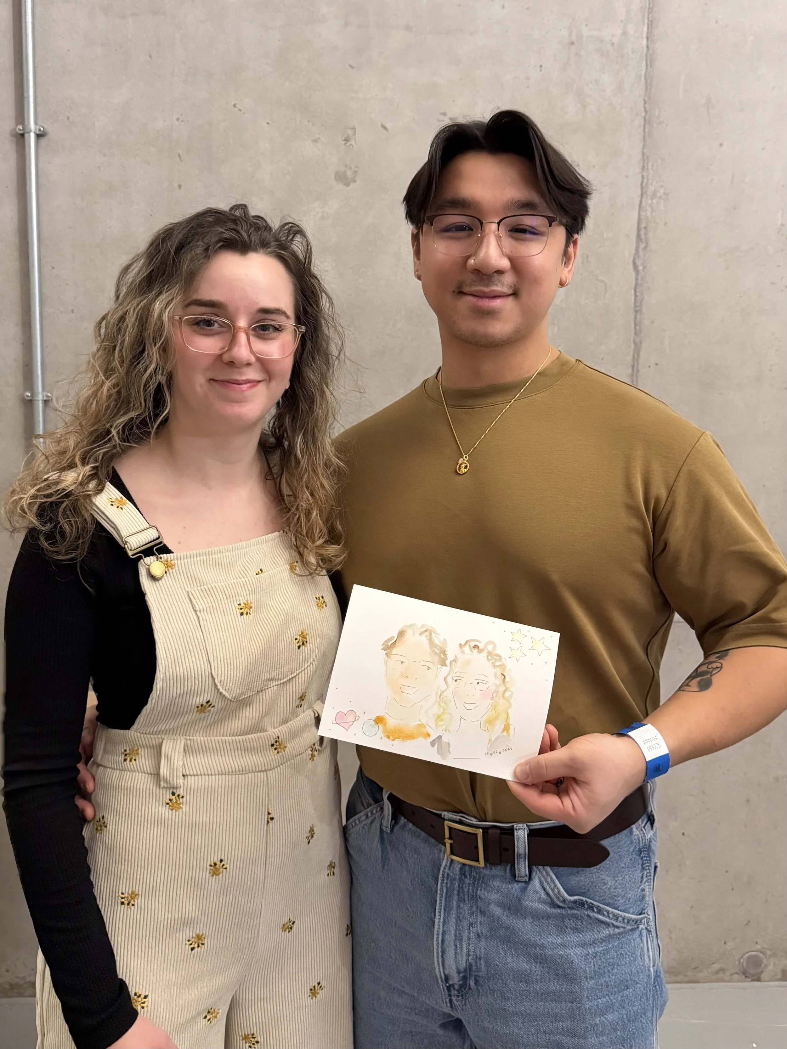 A man and woman standing together against a concrete wall, holding a drawing of two smiling people with stars and a heart. The woman has curly hair, glasses, and a beige apron with yellow flowers. The man has dark hair, glasses, a gold necklace, and 
