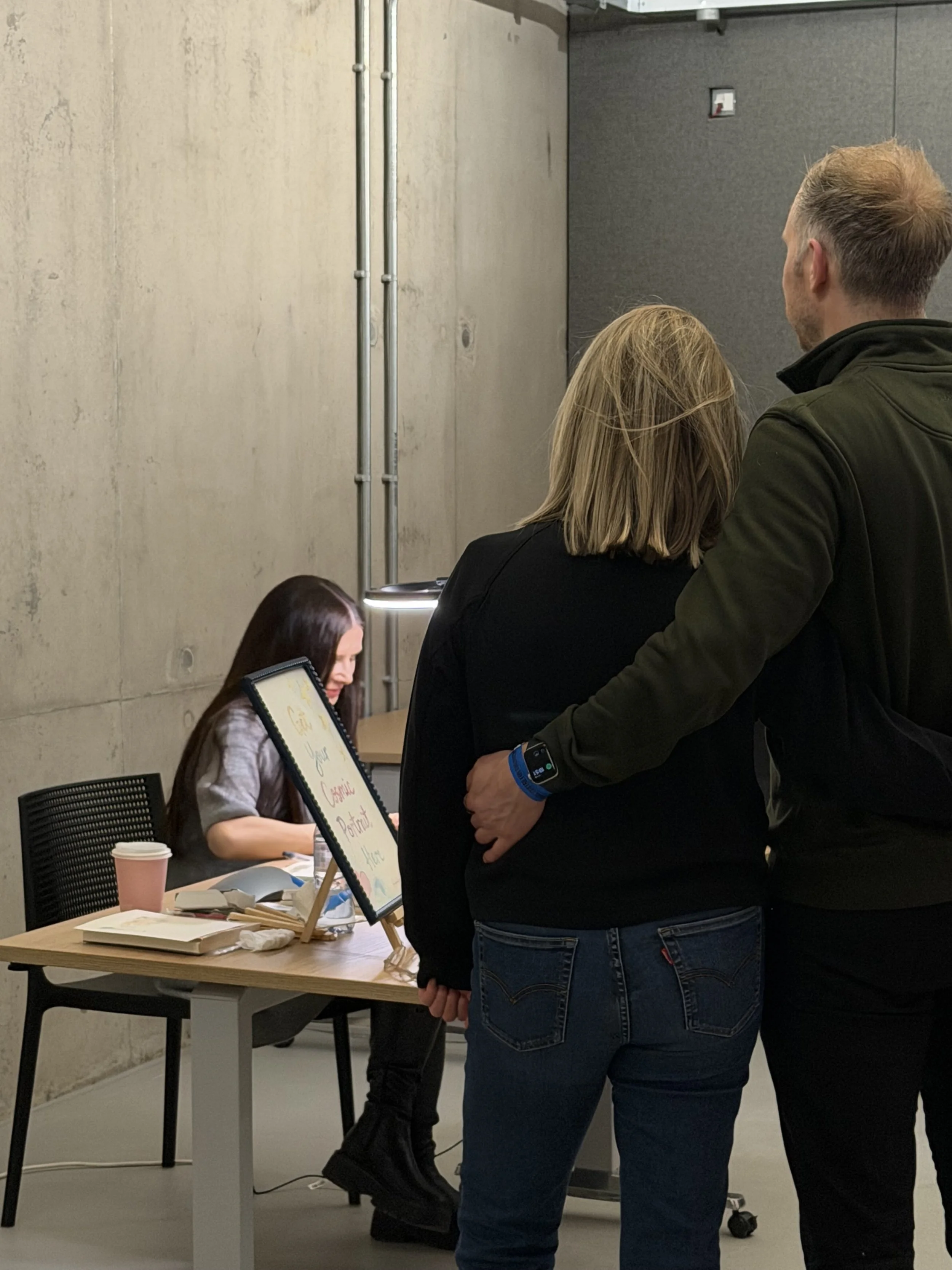 Two people standing in front of a woman sitting at a table in an indoor setting with a concrete wall. The woman is in black boots and appears to be working on a display board. The display board has writing on it, including the words "Serious," "Carin