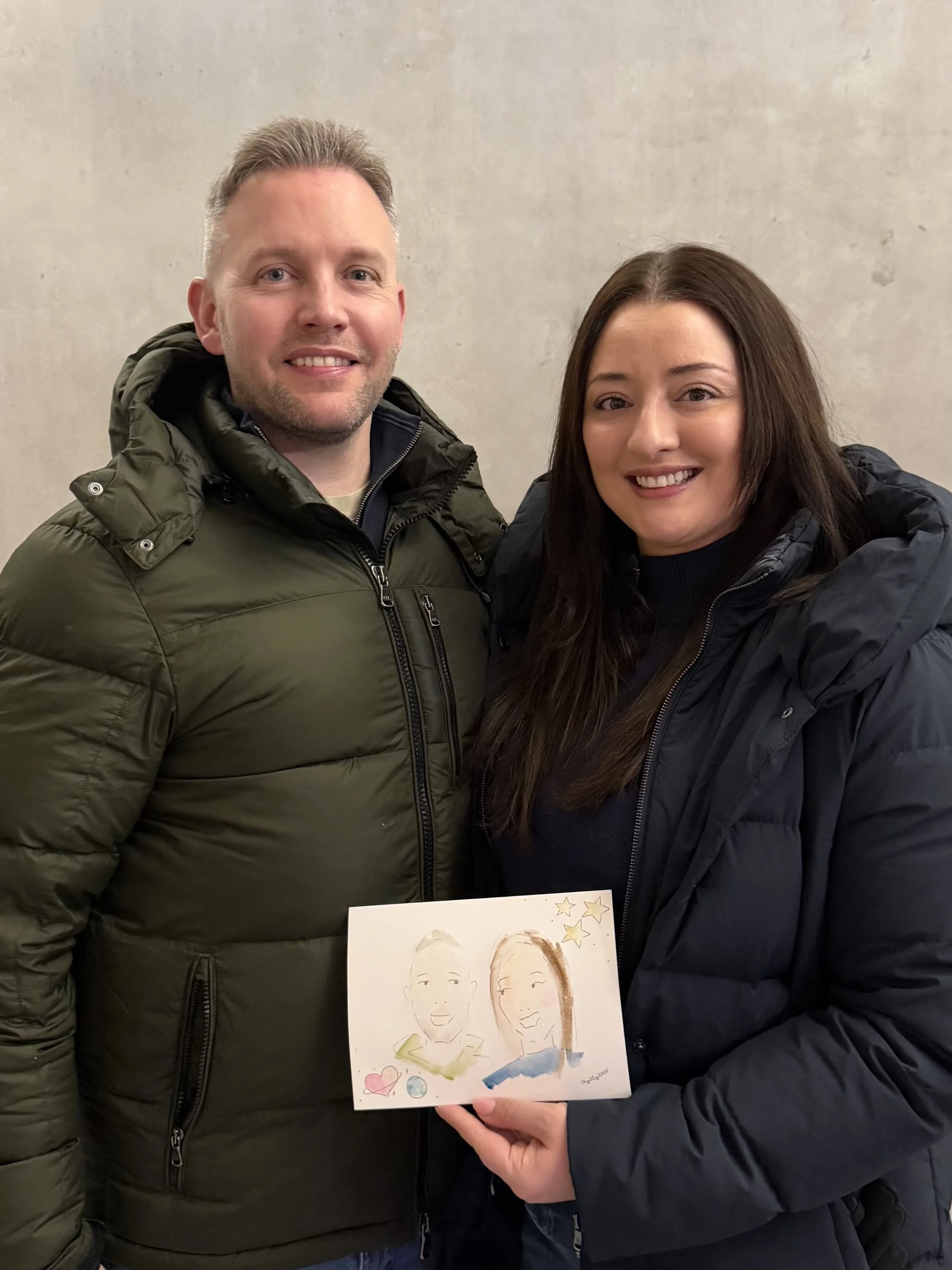 Two people standing together indoors, one holding a drawing of two smiling individuals with colorful decorations, and the other person is wearing a black jacket.