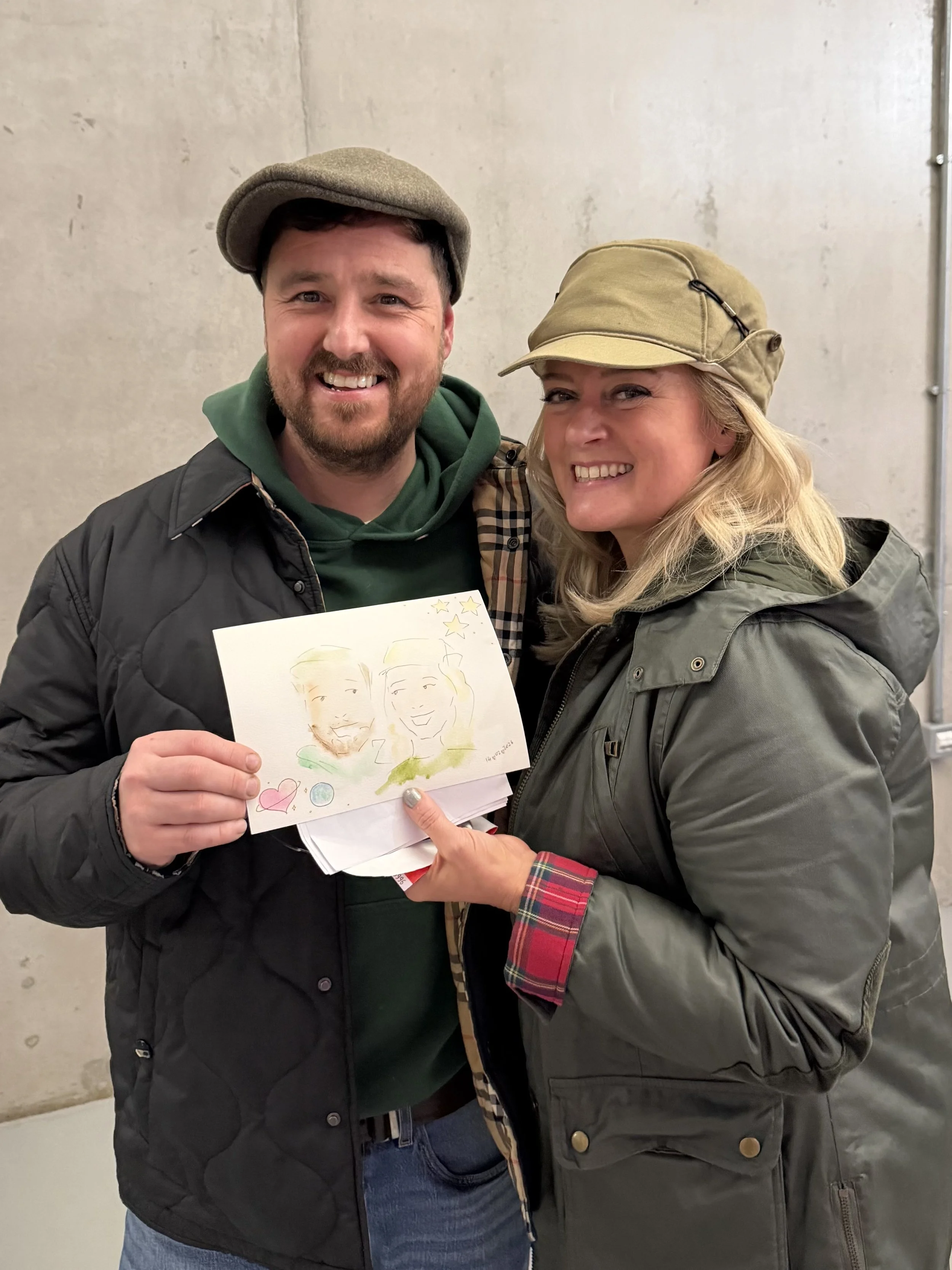 A man and a woman smiling and holding a drawing together. The man is wearing a gray hat and a black jacket. The woman is wearing a tan cap and a gray jacket. They are standing in front of a plain wall.