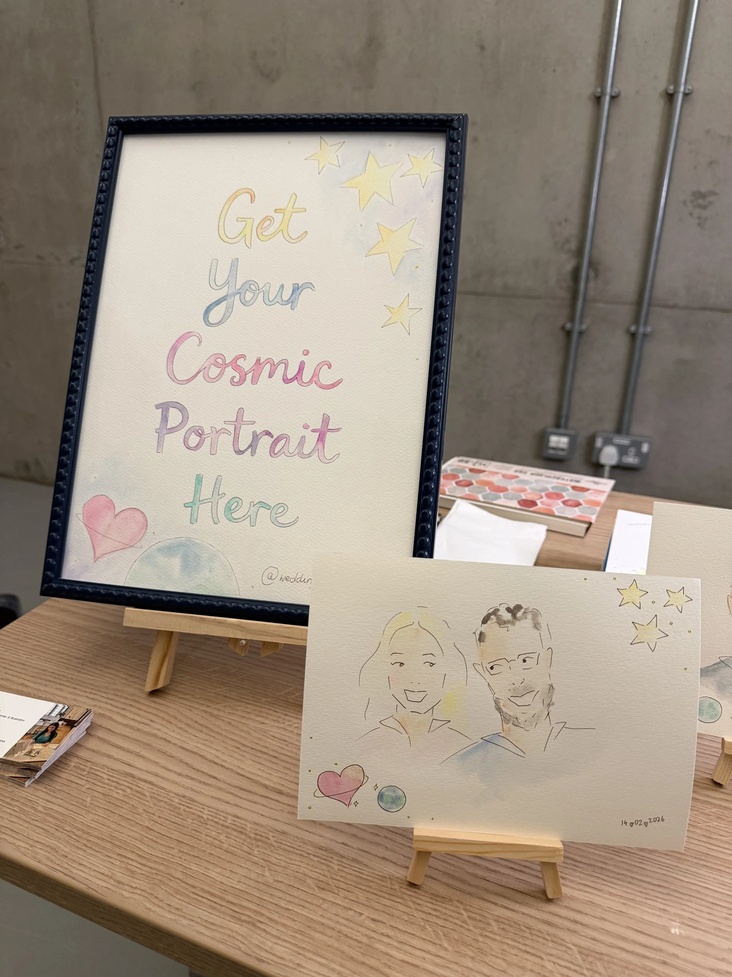A framed sign that reads "Get your Cosmic Portrait Here" and various stars drawings. A hand-drawn illustration of a smiling woman and man, along with hearts and planets, sit on a table in front of the sign.