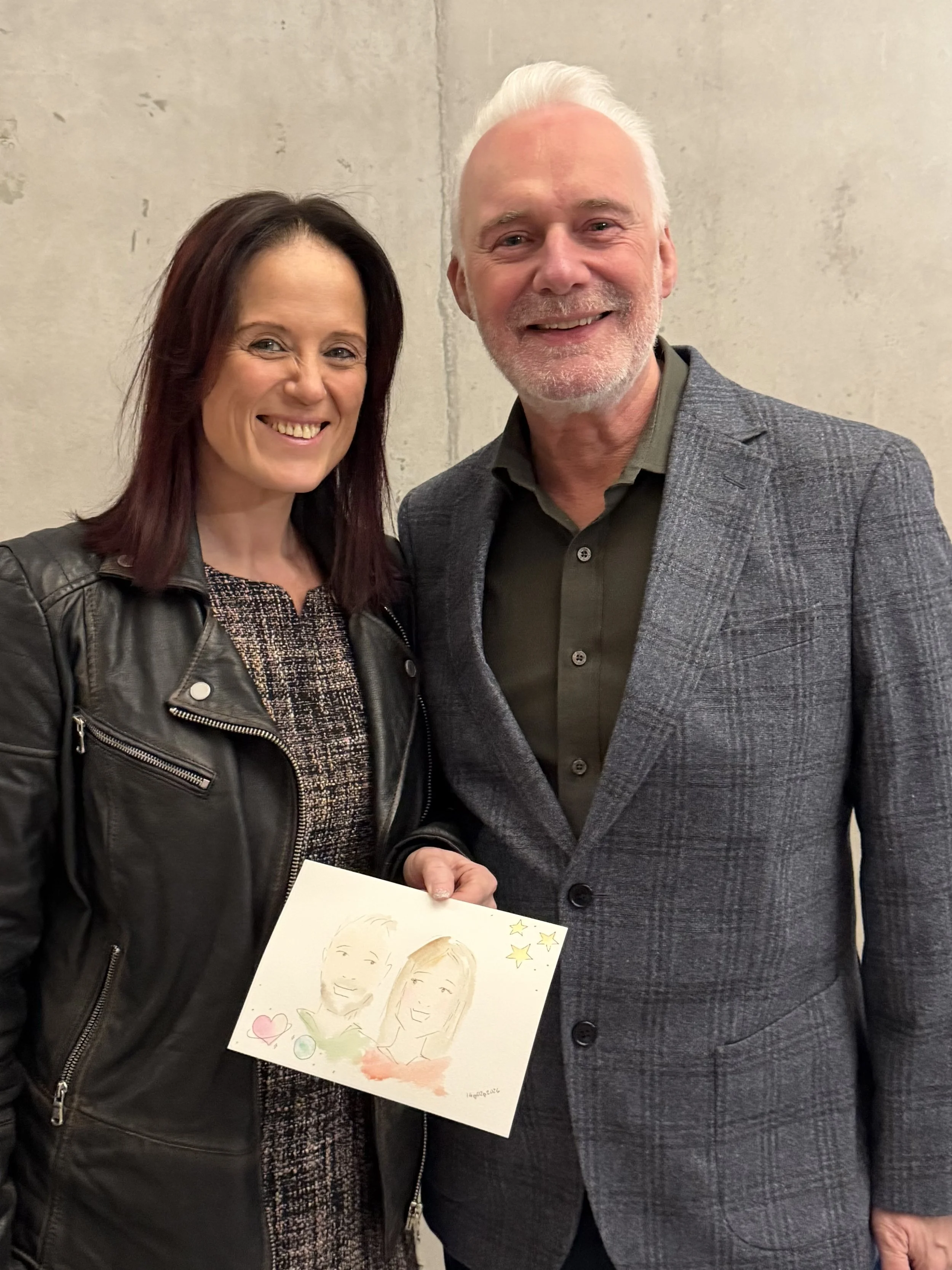 A woman with dark hair and a black leather jacket smiling and holding a colorful caricature drawing of herself and a man. A man with white hair, beard, and a gray checked blazer is smiling beside her.