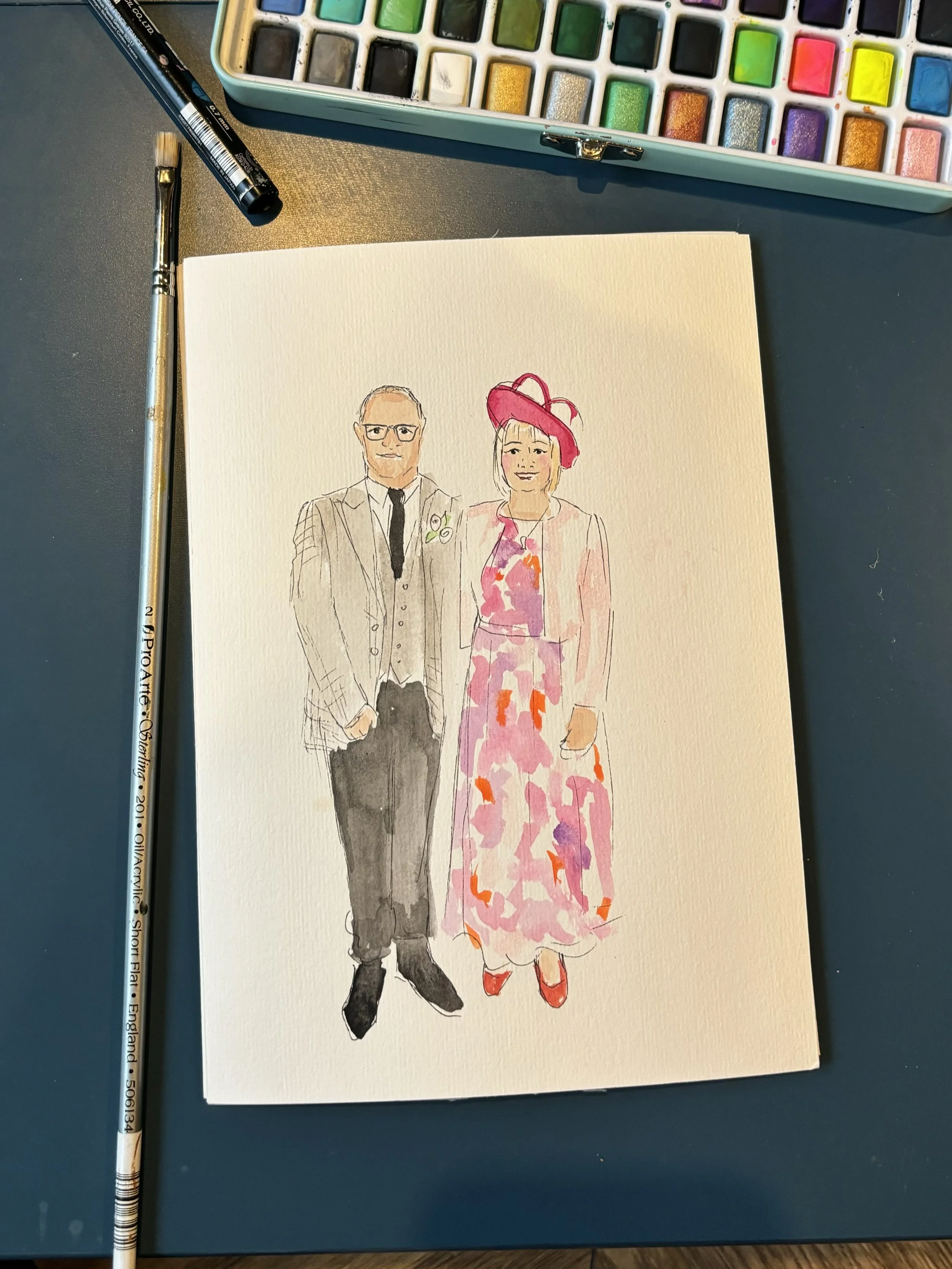 A watercolor illustration of a man and woman standing side by side, with the man dressed in a suit and the woman wearing a colorful dress and a pink hat, on a sheet of watercolor paper with watercolor paints and brushes nearby.
