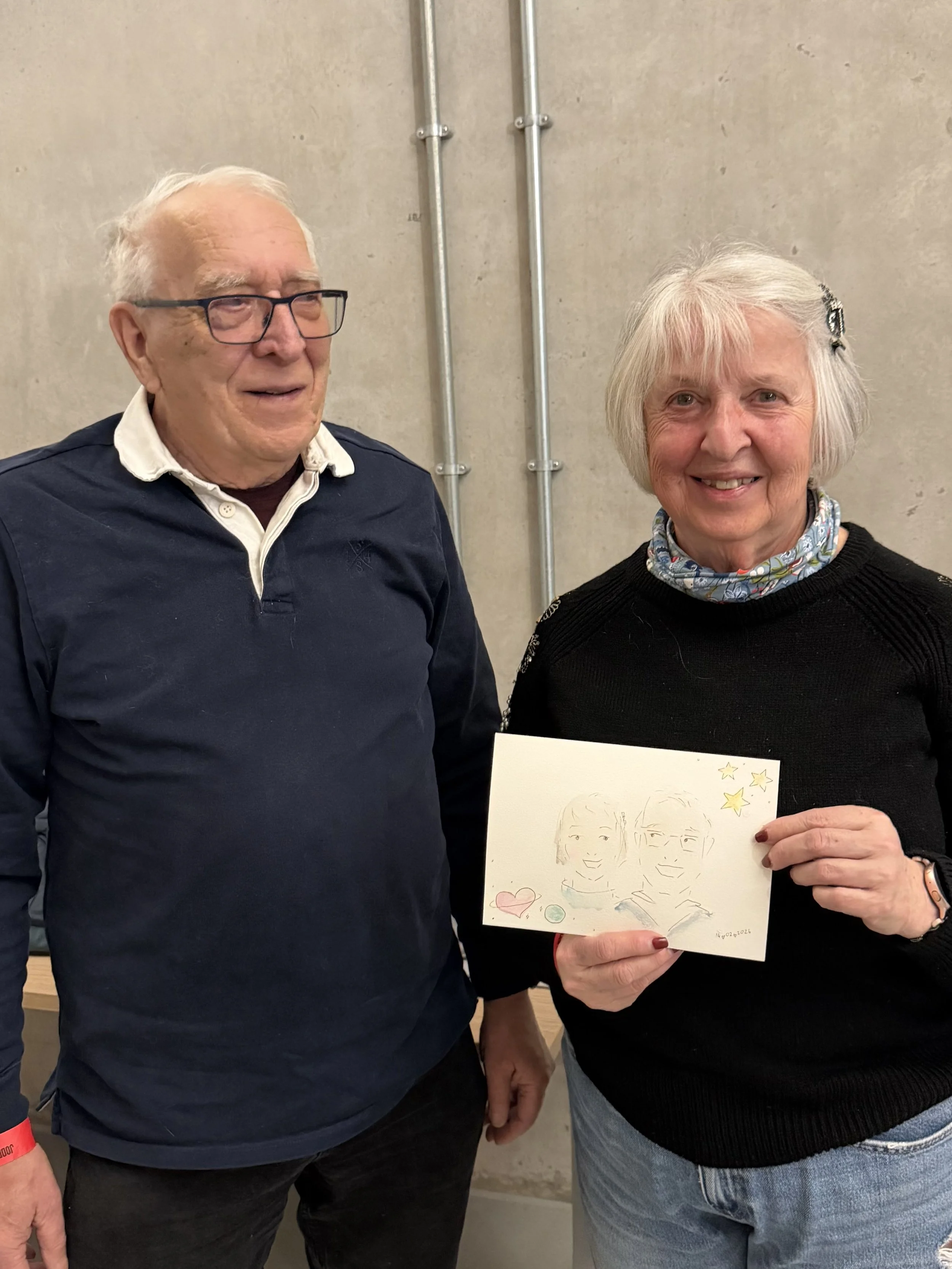 An elderly man and woman standing together, holding a drawing of two smiling faces and stars on a card.