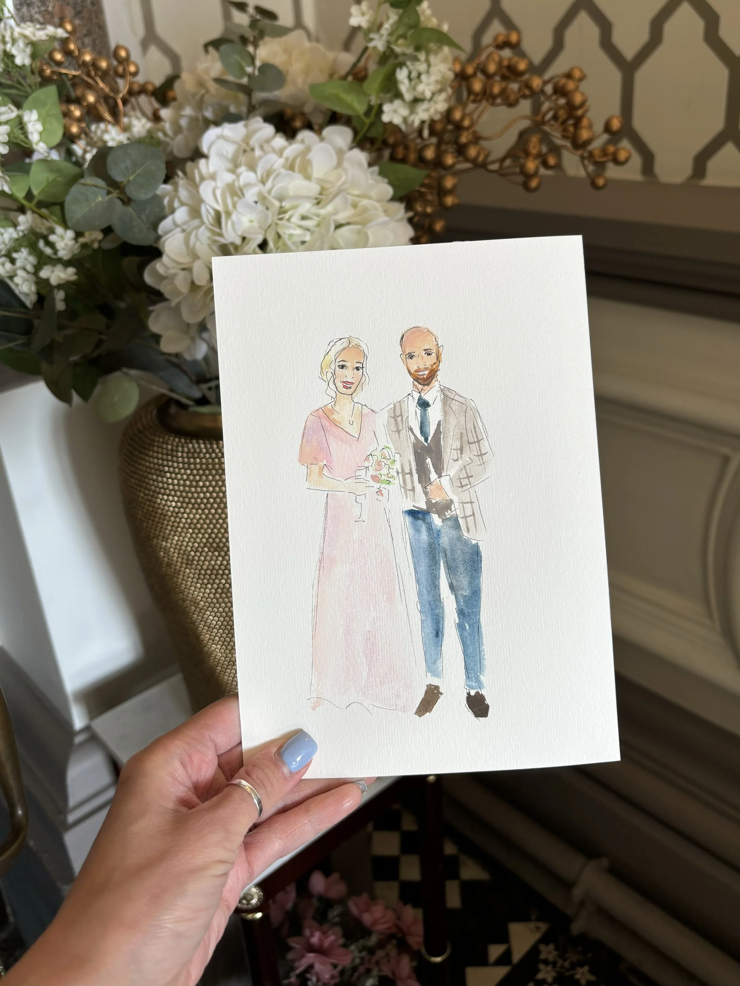 A hand holding a watercolor illustration of a bride and groom, with a lush floral arrangement in the background.