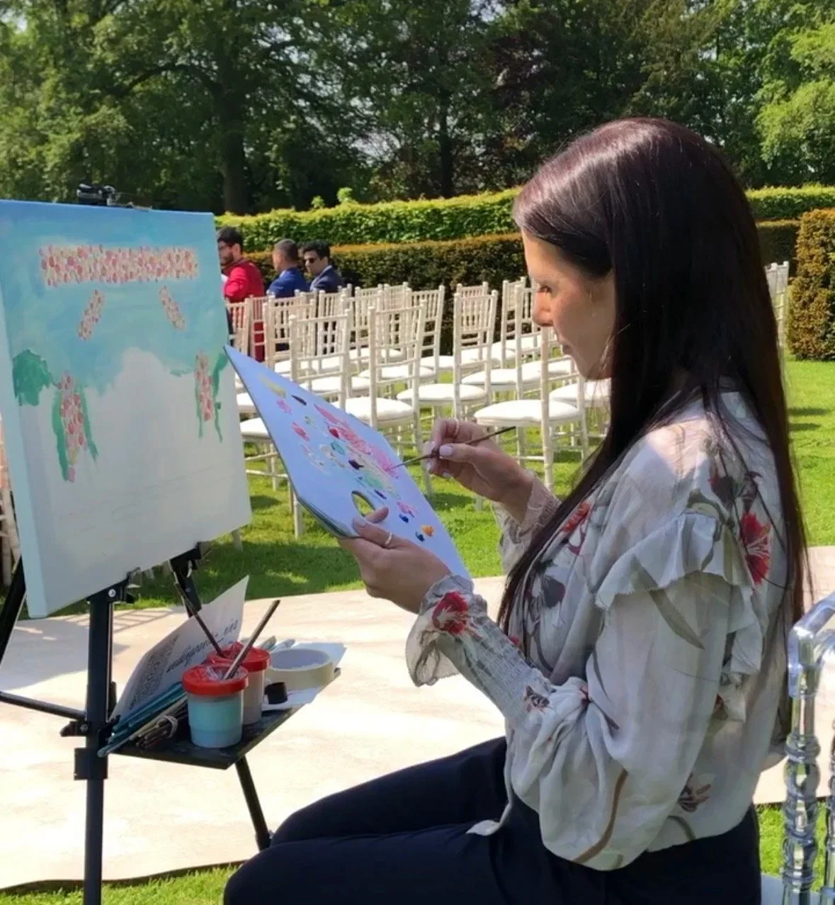 A woman painting a large picture outdoors on a sunny day, with white chairs arranged in rows behind her.
