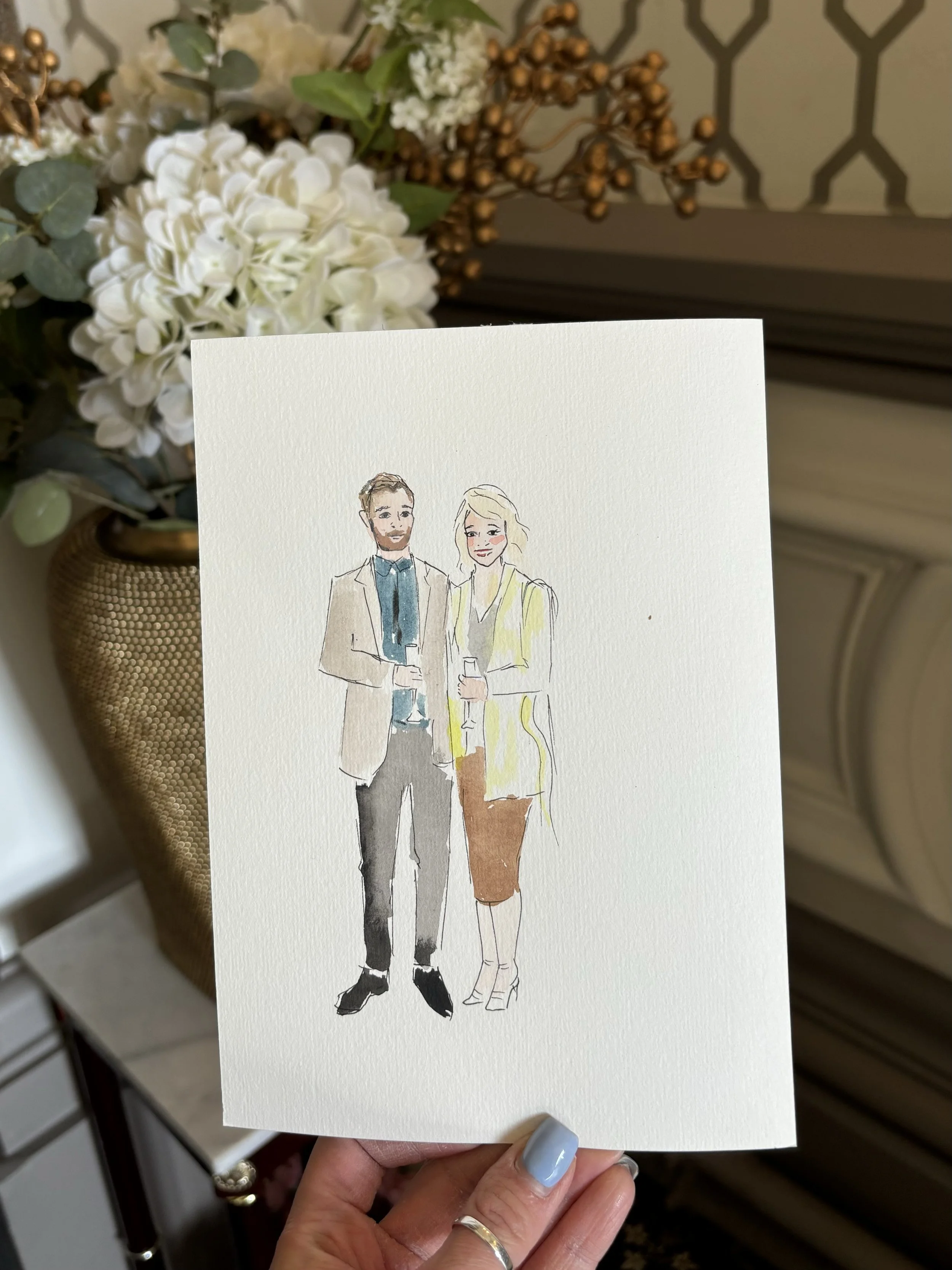Handheld greeting card with a watercolor sketch of a man and woman dressed in formal attire, holding glasses, surrounded by floral arrangements in the background.
