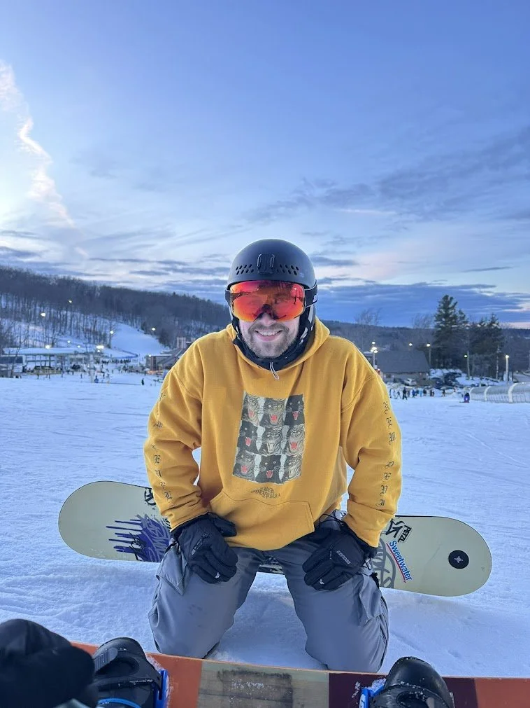 A man in snow gear with a black helmet and red goggles, sitting on snow with a yellow hoodie and snowboard, smiling at a ski resort during early evening with snowy ski slopes and buildings in the background.