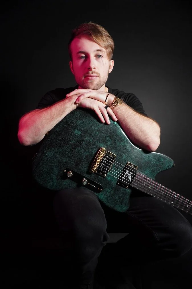 A young man with light brown hair and a beard, wearing a black t-shirt, is sitting with his arms resting on an electric guitar, looking at the camera against a black background.