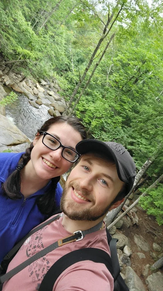 A smiling man and woman taking a selfie outdoors near a rocky stream in a lush green forest.