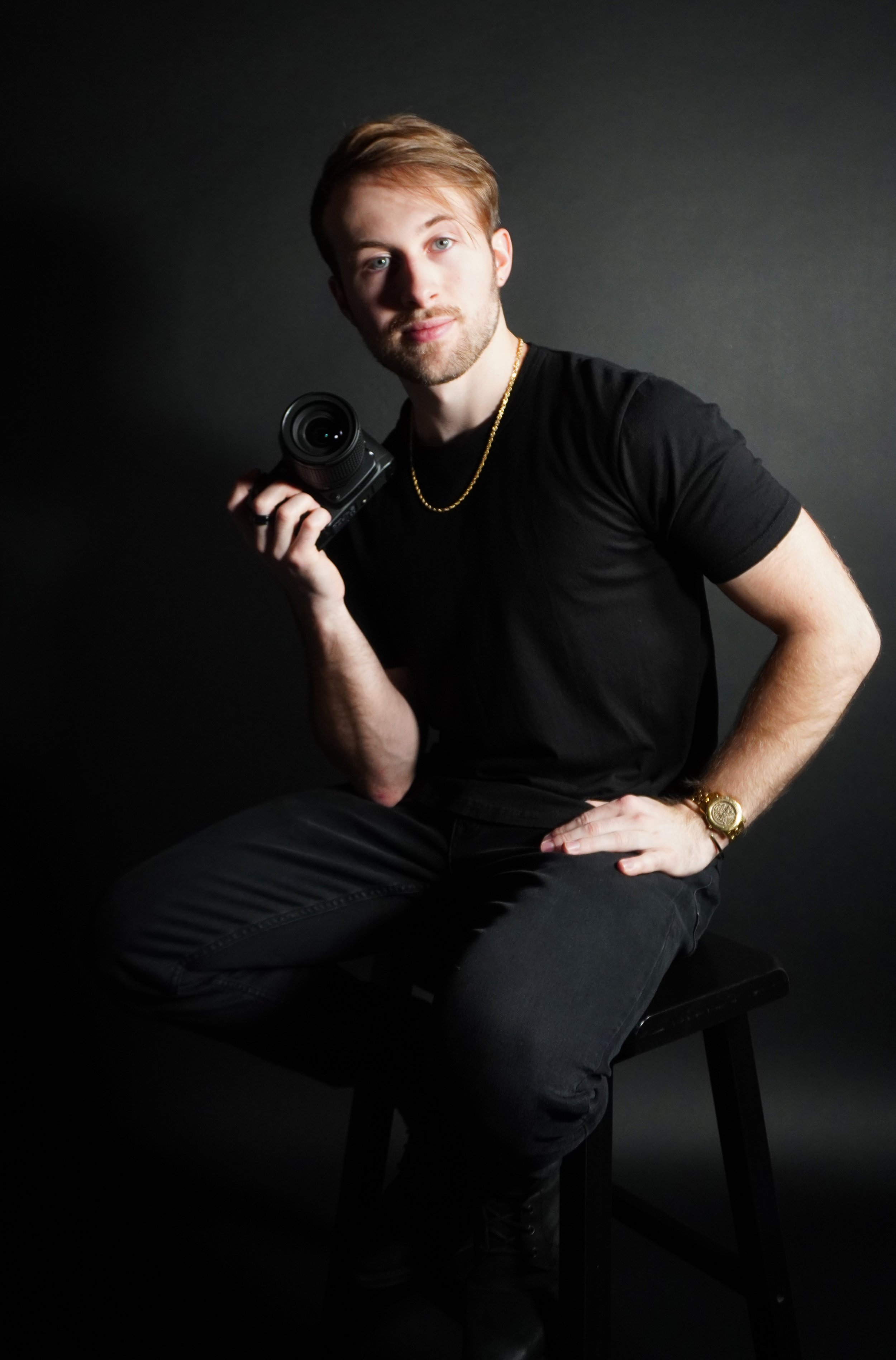 A young man with blond hair and light skin sitting on a black chair against a dark gray background. He is holding a camera in his right hand, resting it on his shoulder, and has his left hand on his thigh. He is wearing a black t-shirt, black pants, a gold chain necklace, a gold watch, and a ring on his right hand.