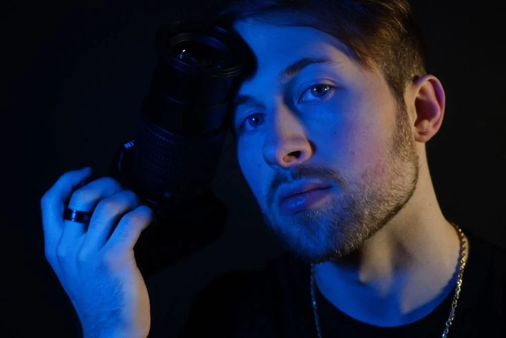 A young man looking into the camera with a serious expression, holding a camera lens close to his face, with blue lighting on his face and dark background.