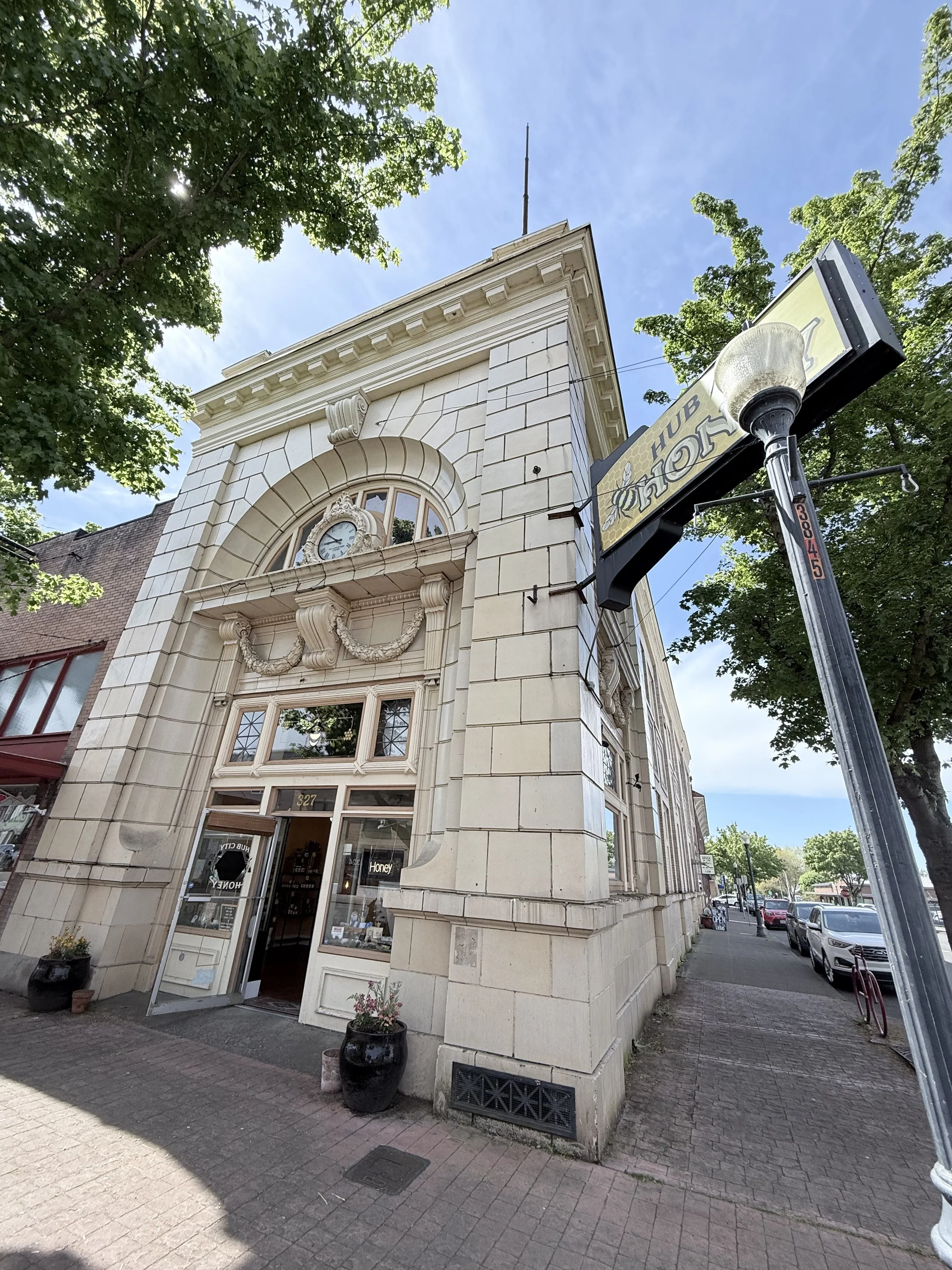 Street view of a beige historic building with a clock above the entrance, decorative trim, and large windows, trees lining the sidewalk, and a sign for a honey shop.