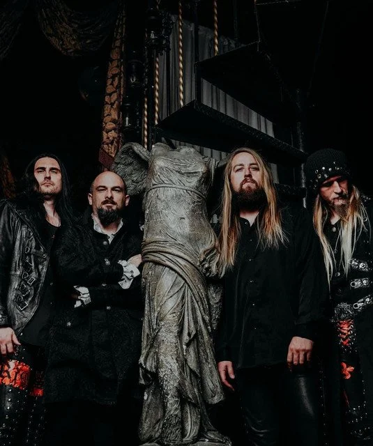 Five men in dark clothing standing behind a stone sculpture of a woman with long hair, in a dimly lit room.