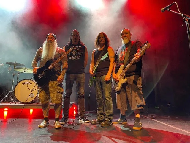 Four musicians standing on a stage with musical equipment, colorful stage lighting, and smoke effects.