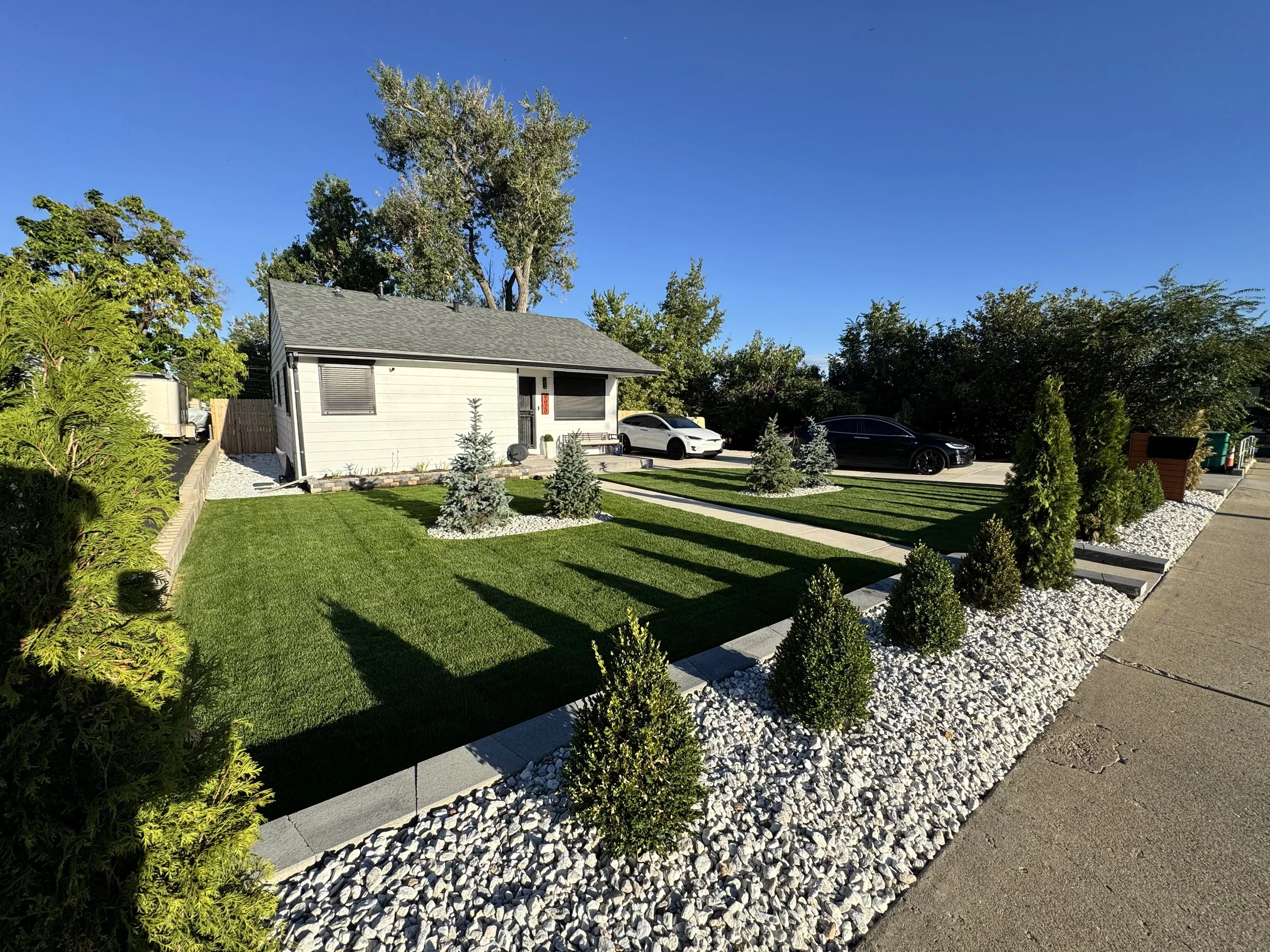 A neatly landscaped front yard with a small grey house, a lawn, small trees, and shrubs, with three cars parked on the driveway, under a clear blue sky.