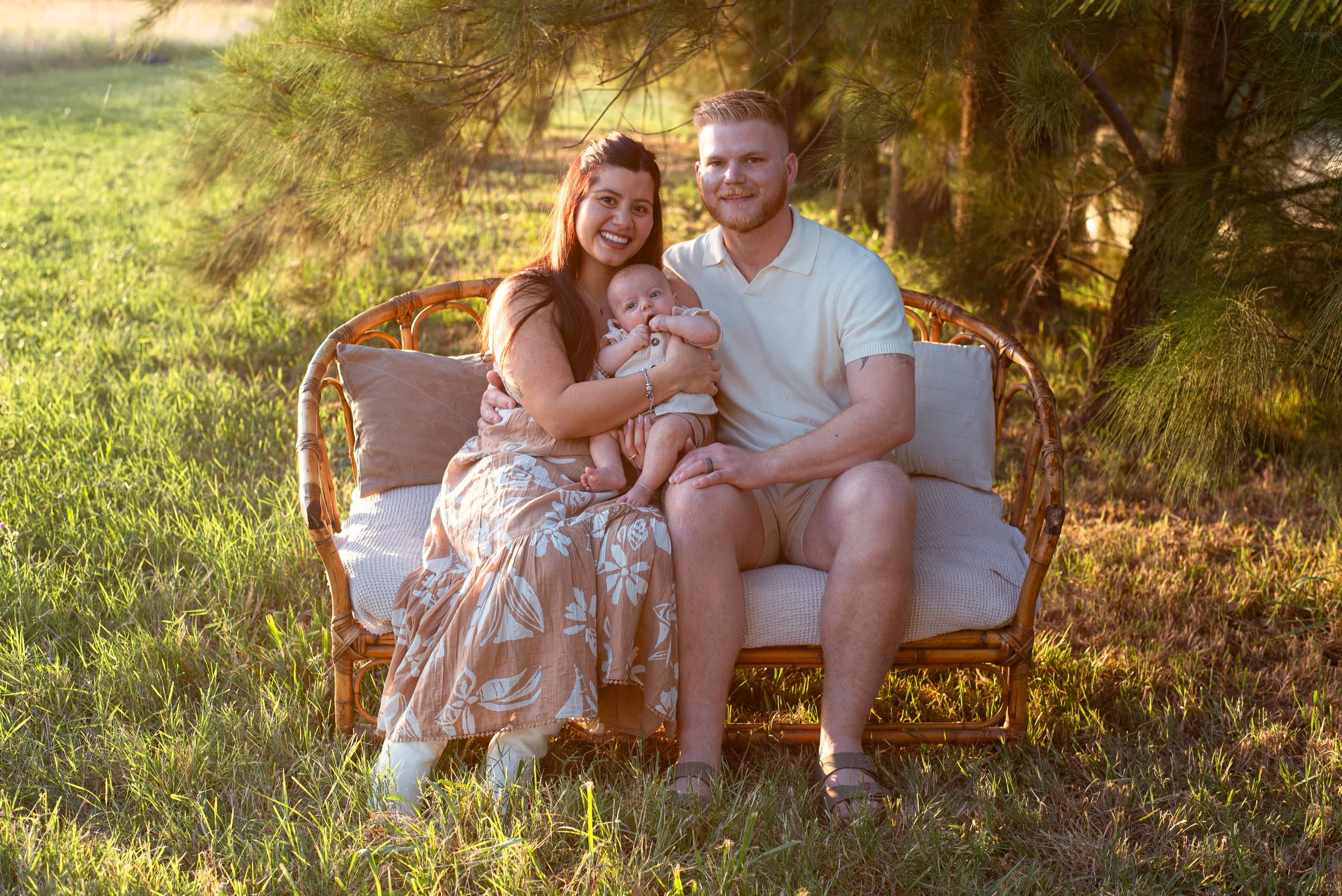 A happy family of three sitting on a wicker loveseat outdoors in a park during sunset. The woman has long reddish-brown hair, wearing a floral skirt and white top, holding a baby. The man has a beard, wearing a white polo shirt and khaki shorts, sitting beside her. The baby is dressed in light-colored clothing, sitting on the woman's lap, and appears to be chewing on their hand. The background features green grass and trees with golden sunlight filtering through.