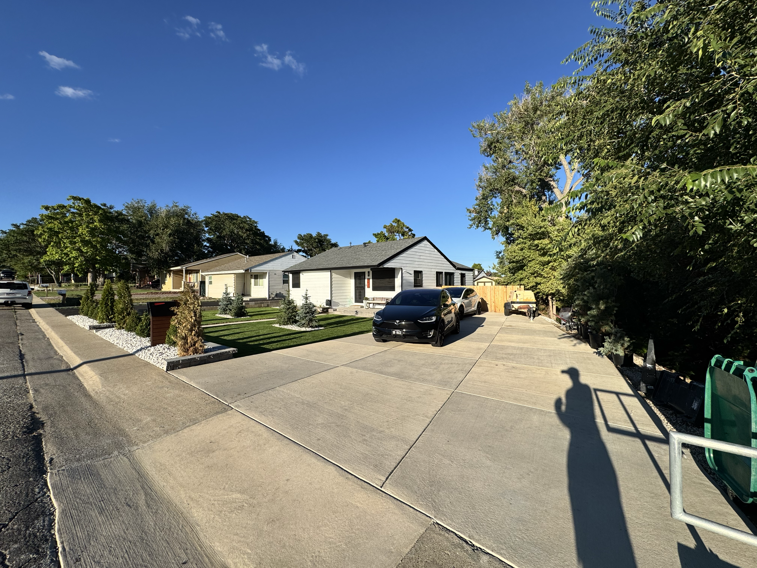 A residential street with new houses, parked cars, and a sidewalk. The sky is blue with some clouds, and the sun casts shadows on the driveway and street.