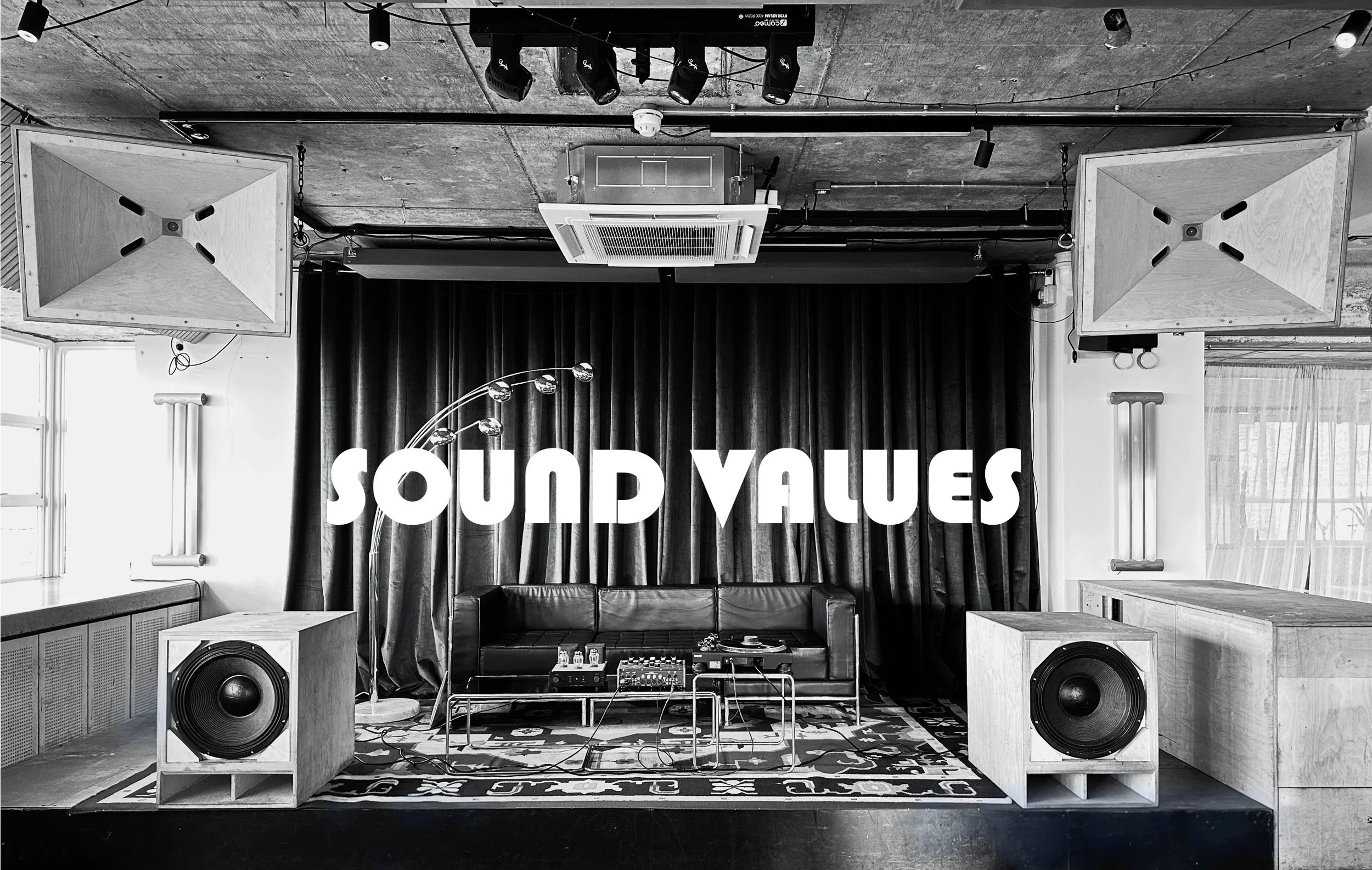 A stage with sound equipment, a black leather sofa, and a large sign that says 'Sound Values' in white block letters. The stage has two large speakers, a patterned rug, and a black curtain backdrop. There are decorative items and lighting fixtures above the stage.