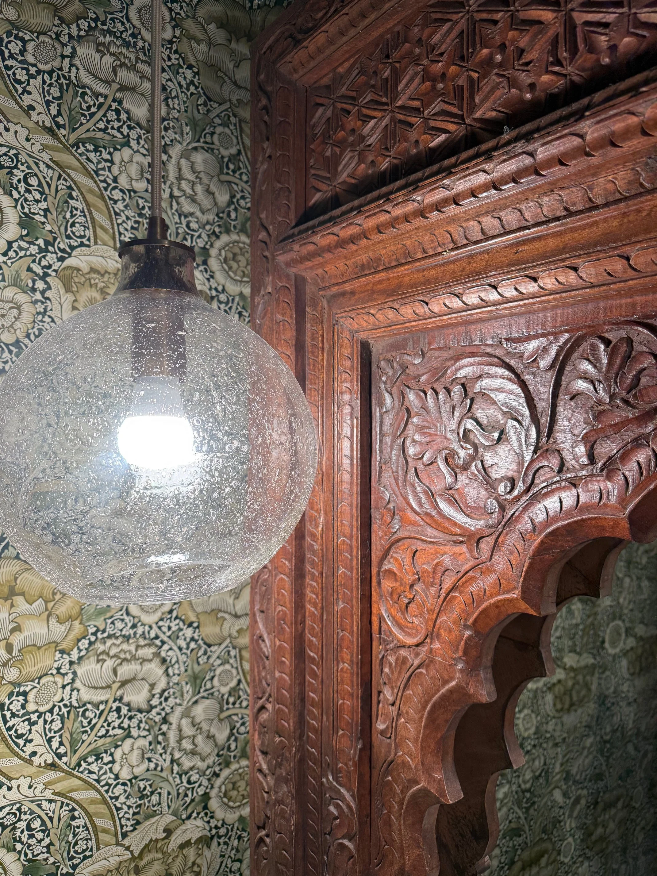 Close-up of a clear, textured glass pendant light hanging next to intricately carved dark wood paneling, with floral-patterned wallpaper in the background.