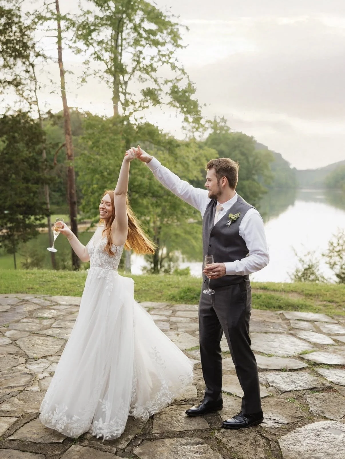 A bride and groom celebrating outdoors by a river, dancing and holding glasses of champagne, with trees and a cloudy sky in the background.