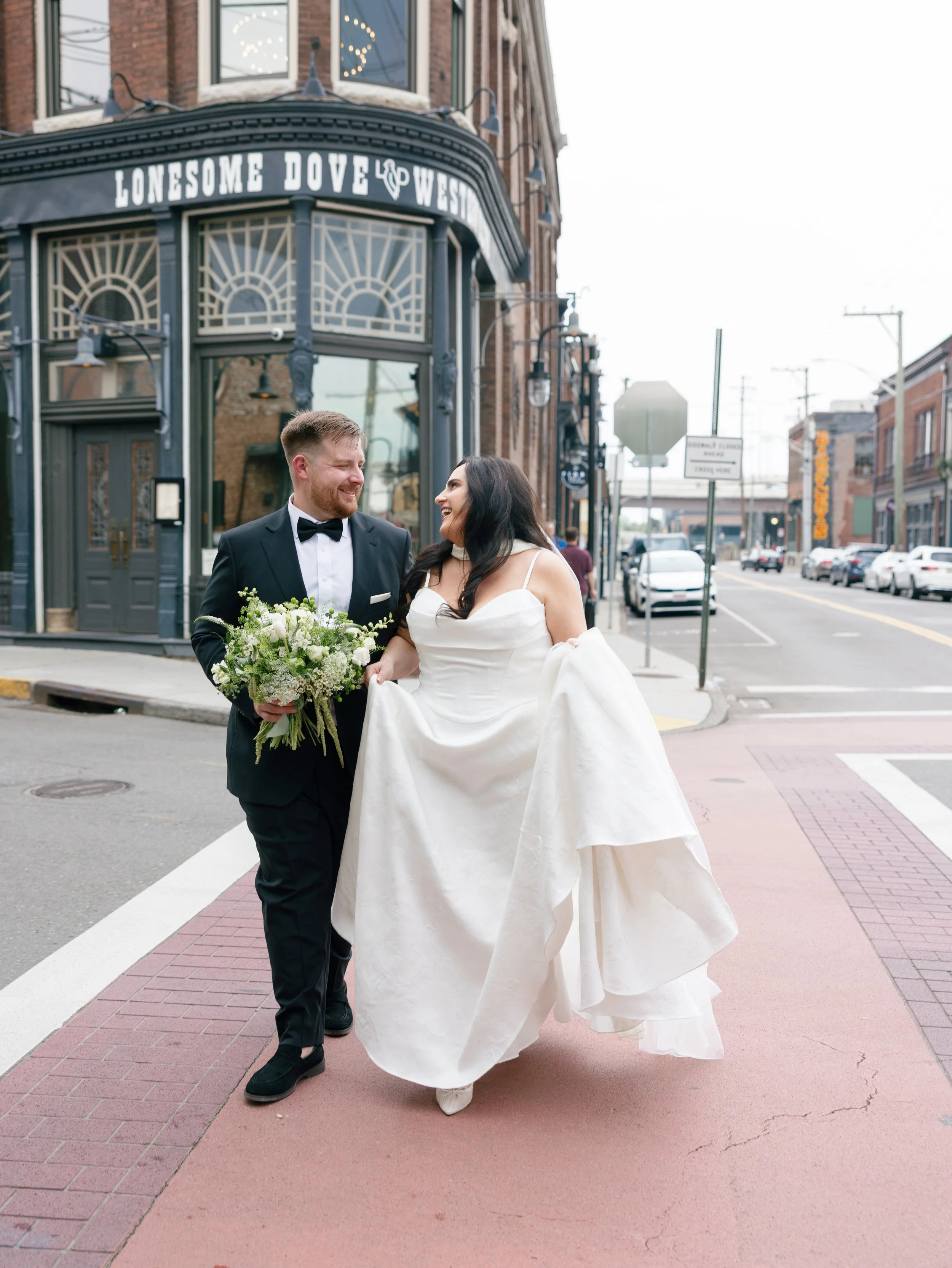 A bride and groom walking on a city street, smiling at each other. The bride is wearing a white wedding dress, and the groom is in a black tuxedo. She is holding a bouquet of white and green flowers. The background features a building with a sign that says "Lonesome Dove West" and storefront windows.
