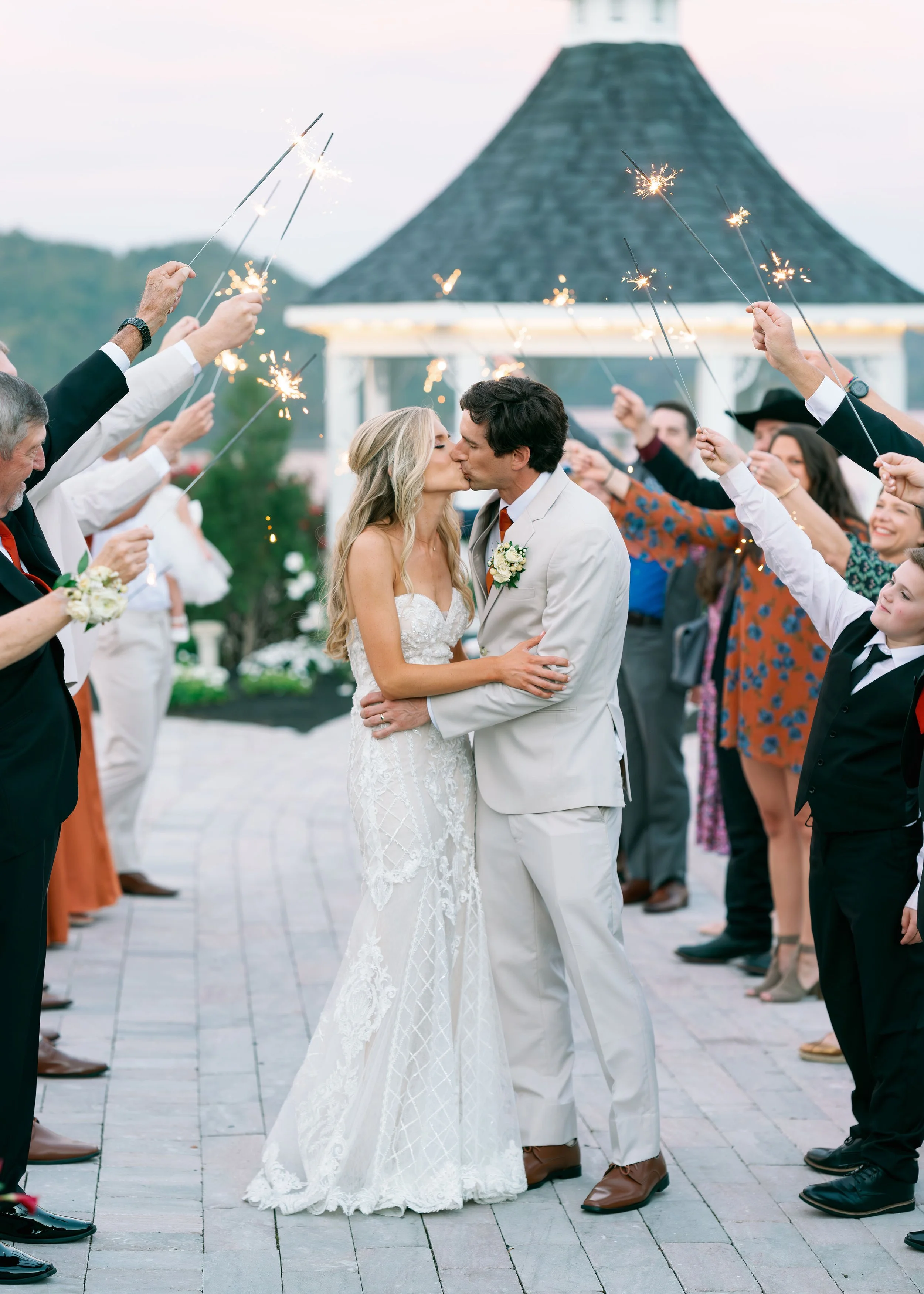 A bride and groom kiss in the center as guests around them hold sparklers. The setting appears to be outdoors near a pavilion with a conical roof.